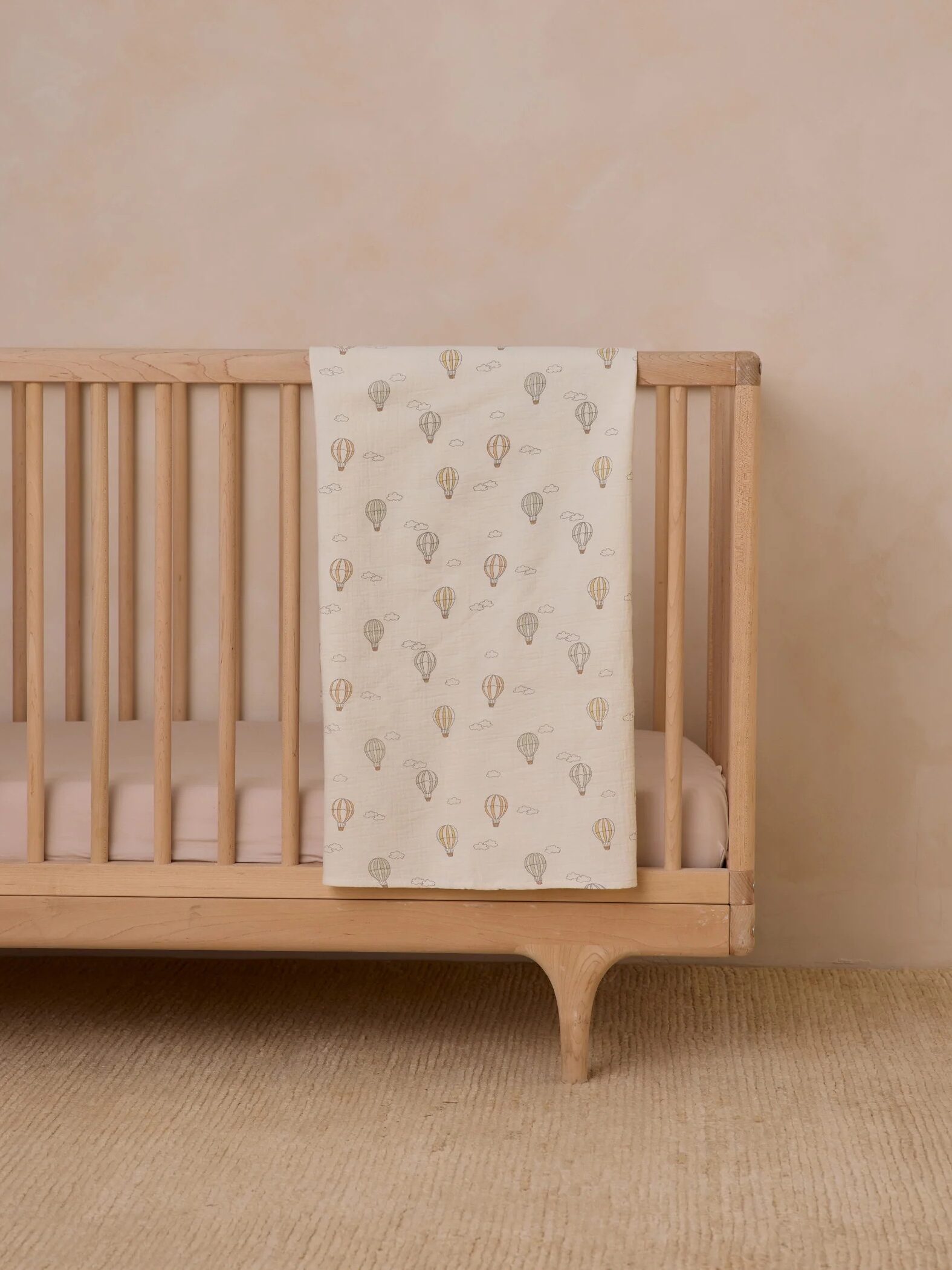 A light wooden crib with a pink mattress and a cream blanket decorated with hot air balloon patterns draped over the side, set on a beige carpet.