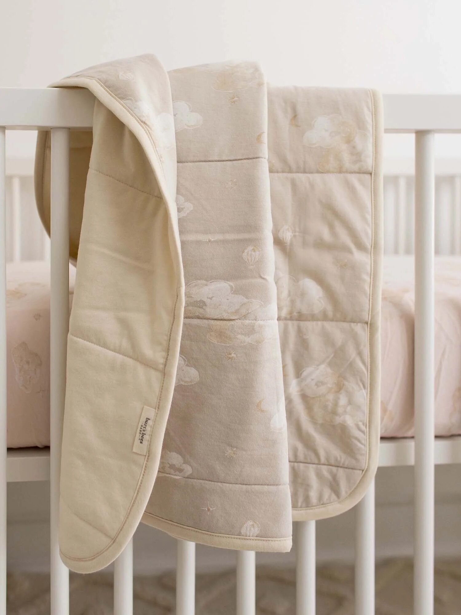 A beige and cream quilted baby blanket with a cloud pattern draped over the side of a white crib with a matching fitted sheet.