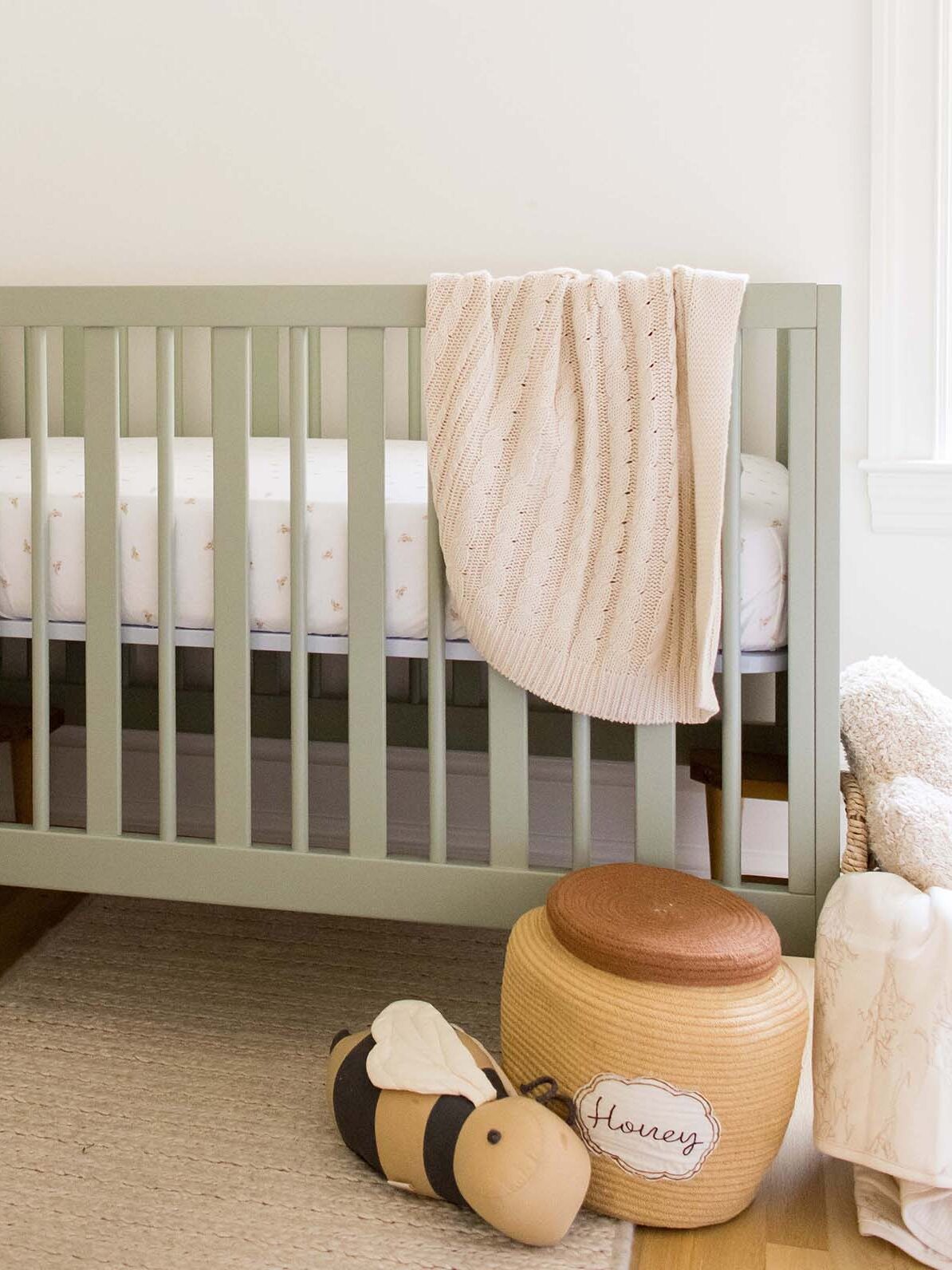 Green baby crib with a cream knit blanket draped over the side, a bee-shaped toy, a basket labeled "Honey," and woven baskets on a light rug in a bright room.