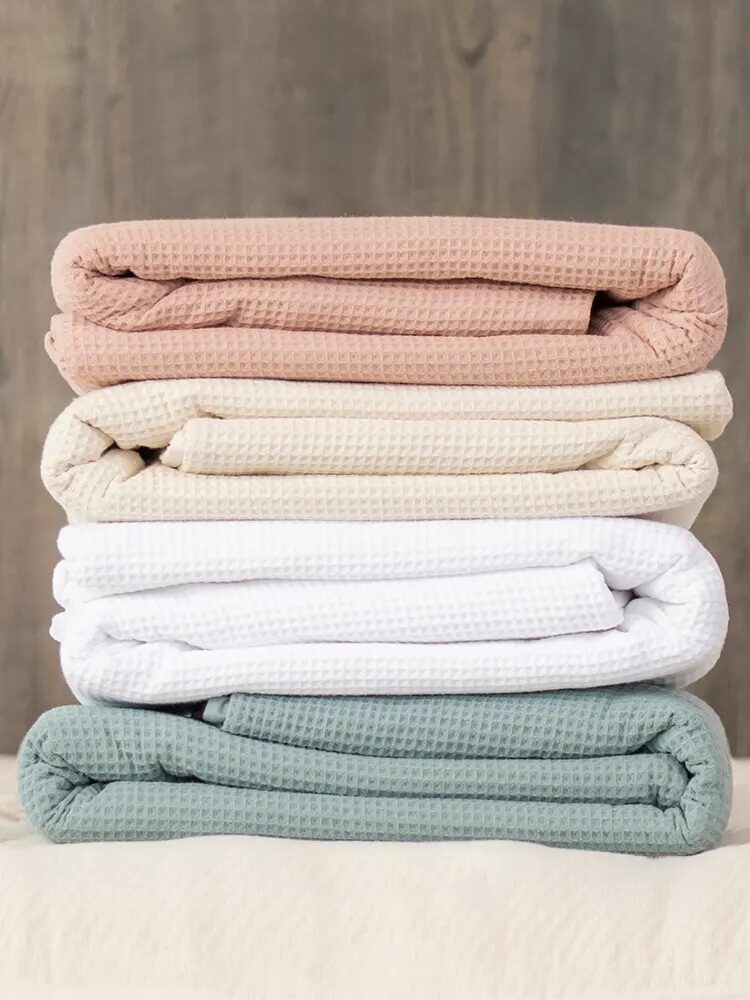 Four folded waffle-texture blankets in pink, beige, white, and green are stacked on a surface with a wood-textured background.