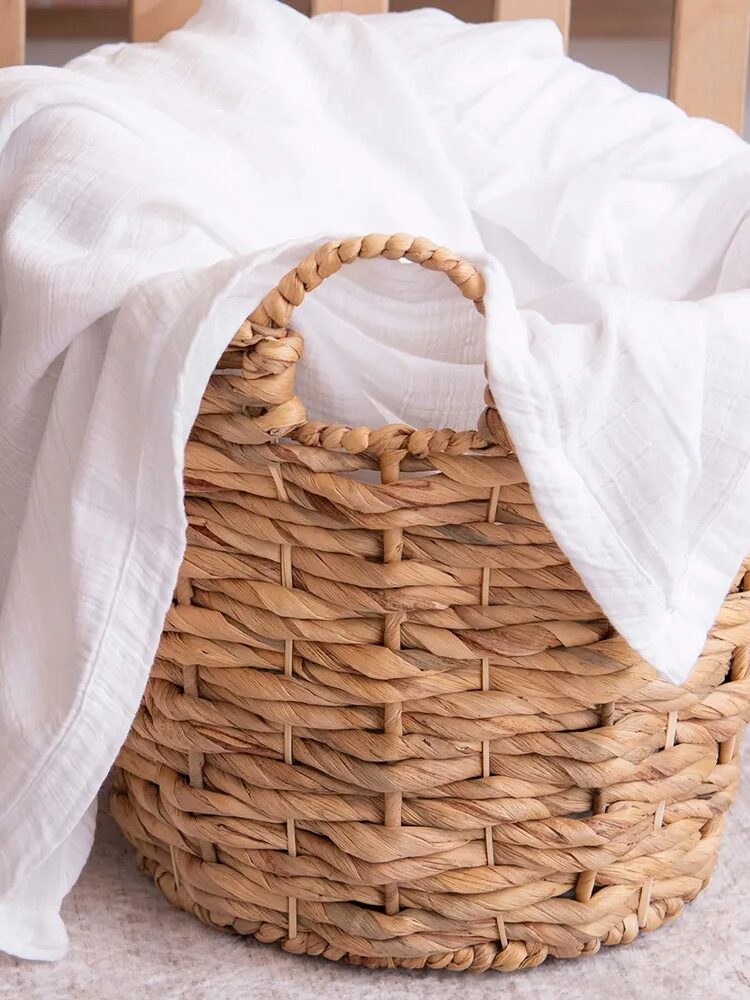 A woven wicker basket with a white cloth draped over its edge sits on a light-colored rug near wooden slats.