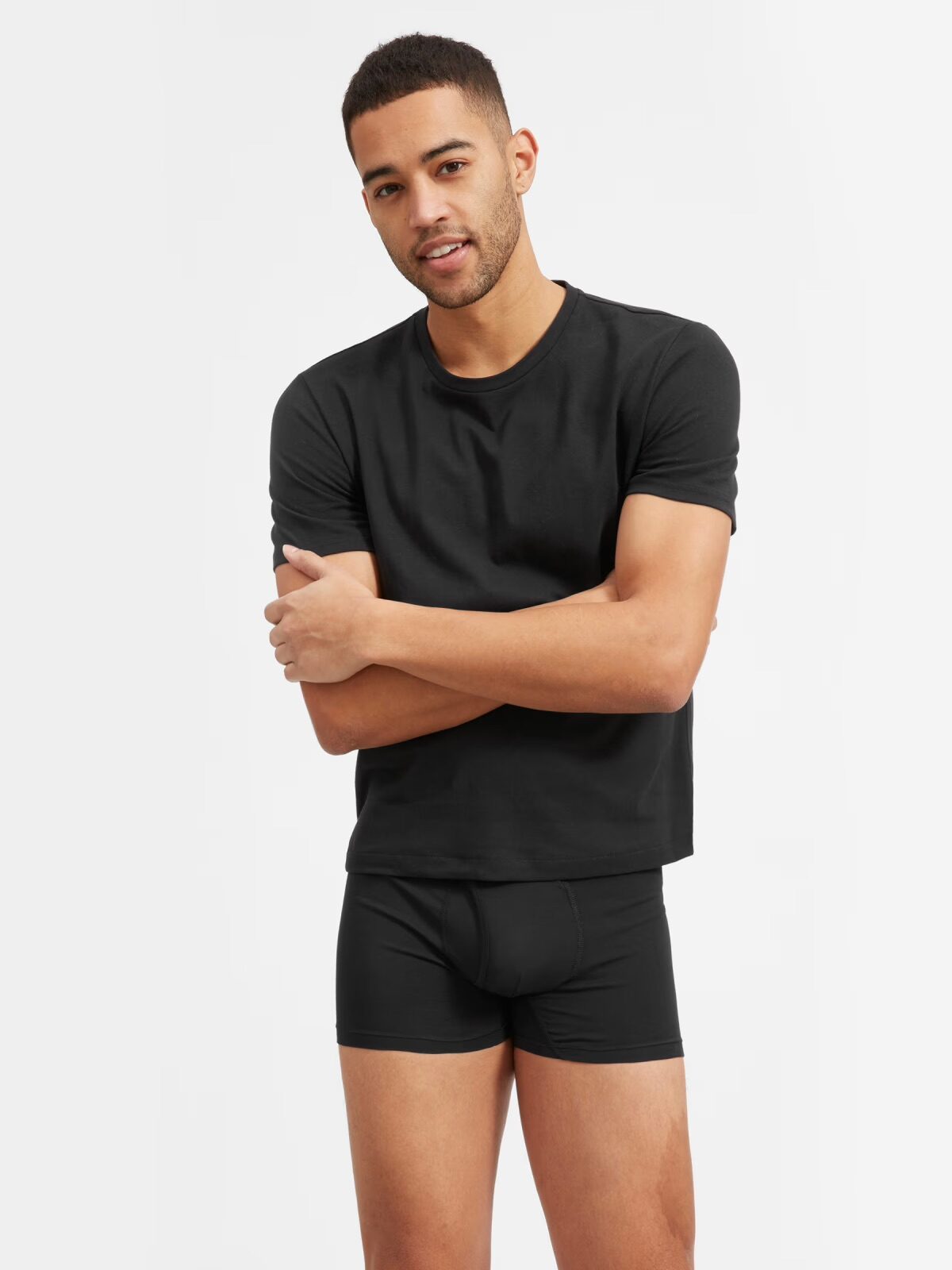 A man stands against a plain background wearing a black t-shirt and black boxer briefs, with his arms loosely crossed and a neutral expression.