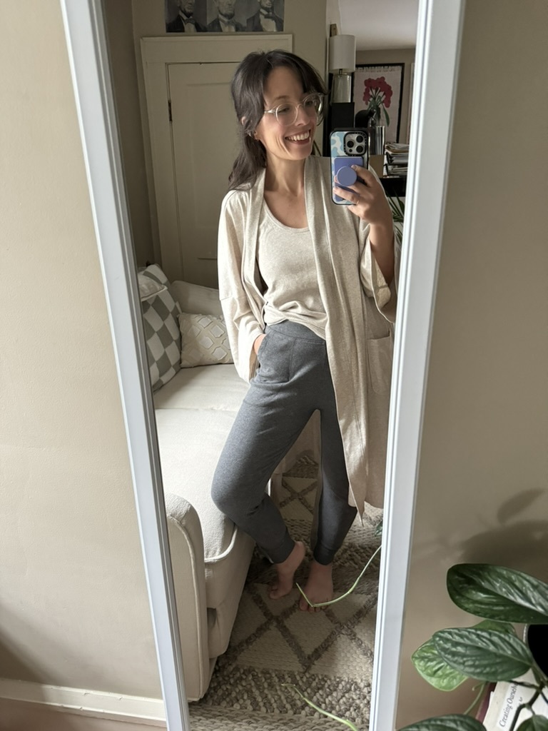 A woman wearing glasses, a beige top, a light cardigan, and gray pants stands barefoot in front of a mirror, smiling and taking a selfie in a living room.