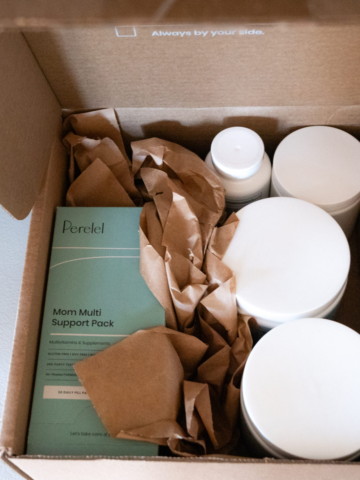 An open cardboard box containing several white plastic containers, a green Perel Mom Multi Support Pack prenatal vitamin, and brown packing paper.