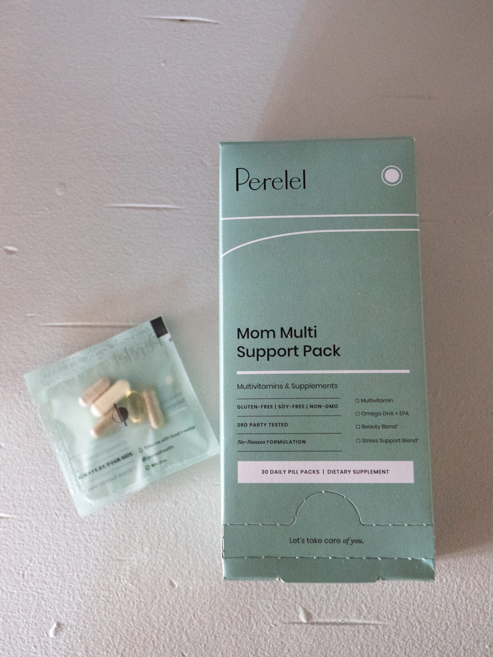 A Perelēl Mom Multi Support Pack box and a small packet of assorted prenatal vitamin pills are placed on a light-colored surface.