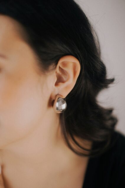Close-up of a person's ear wearing a chunky, silver, geometric-shaped earring; the person has dark hair and light skin.
