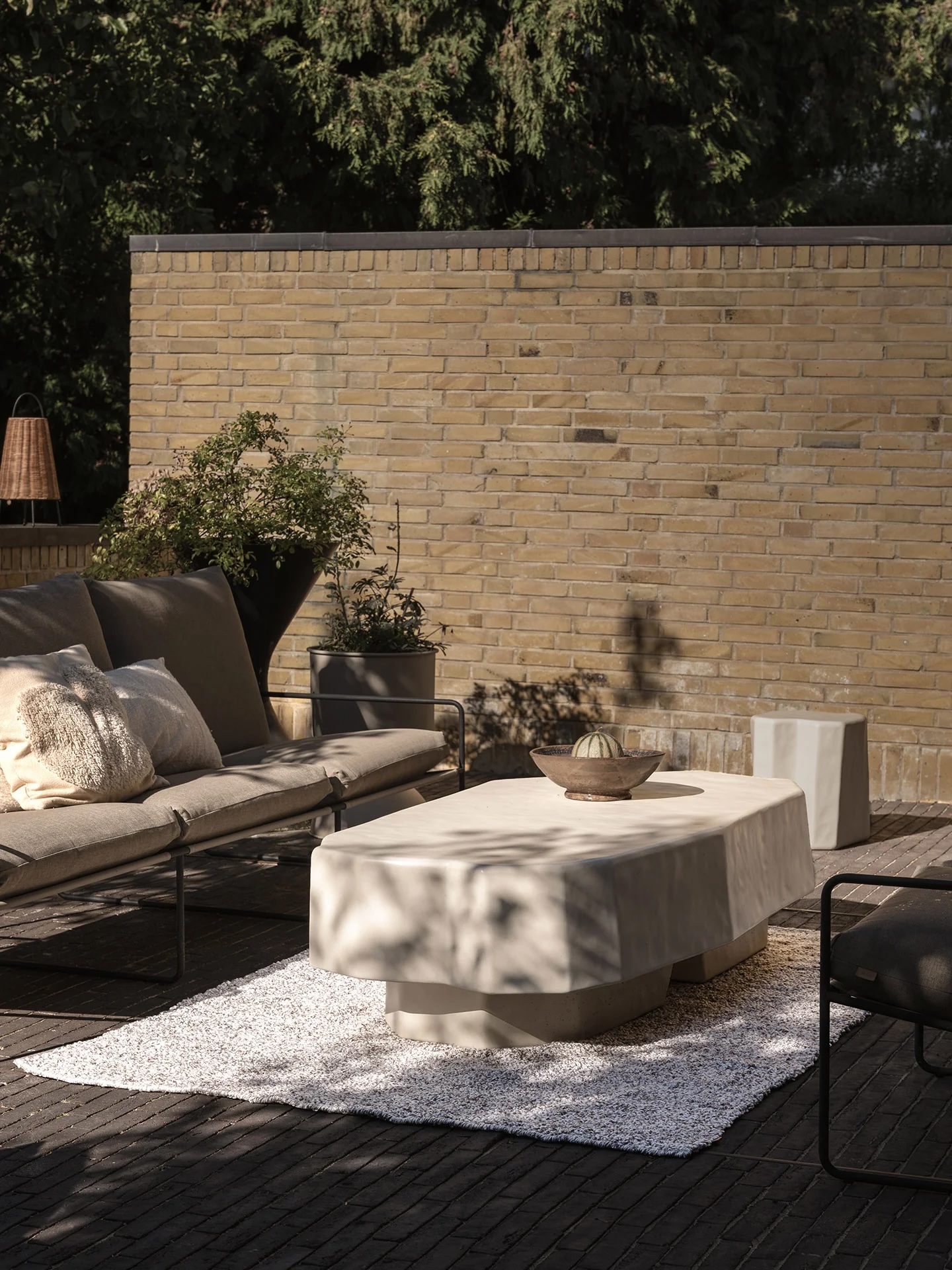 A modern outdoor patio with a beige sofa, neutral pillows, a rectangular coffee table on a white rug, and potted plants against a brick wall.