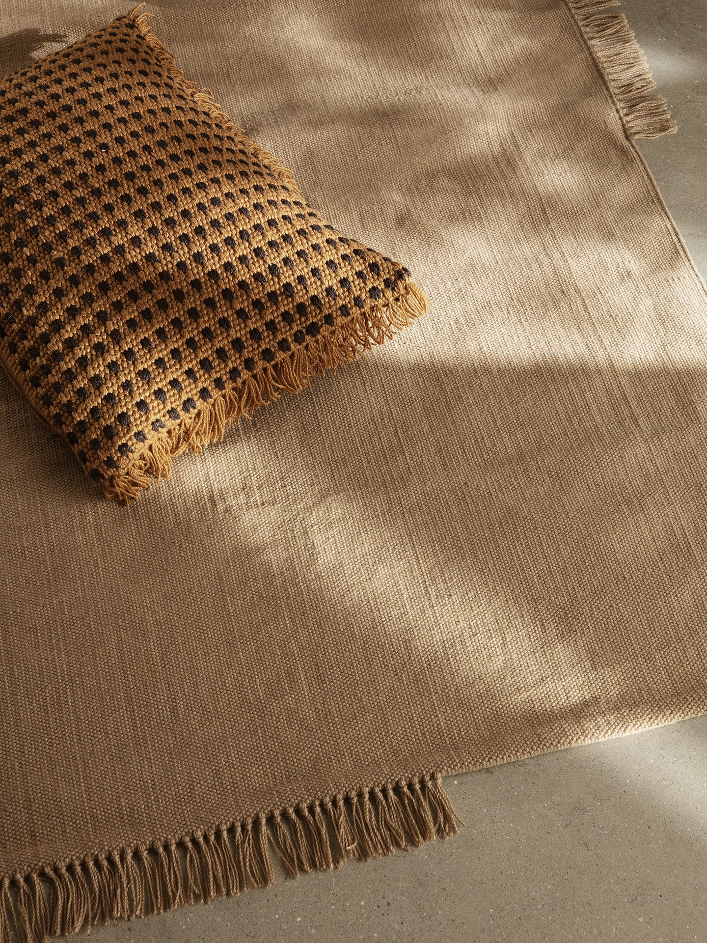 A brown woven pillow with tasseled edges rests on a beige textured rug with fringe on a smooth floor, illuminated by natural sunlight.