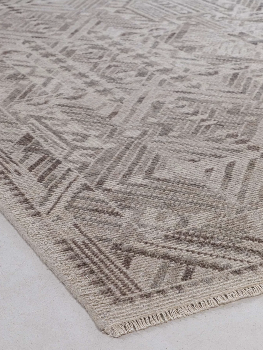 A beige and brown area rug with a geometric pattern and fringed edge on a light-colored floor.
