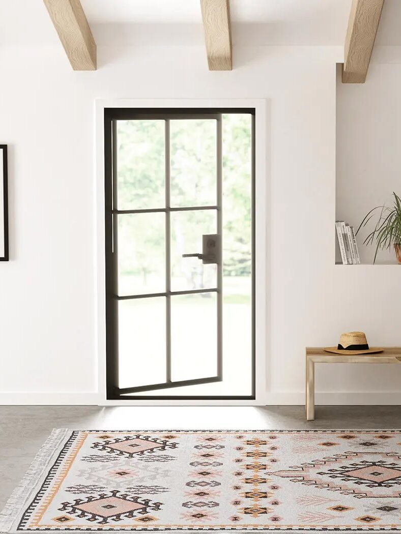 A modern entryway with a black-framed glass door, a patterned rug, a wooden bench with a hat and book, a potted plant, and wall art.