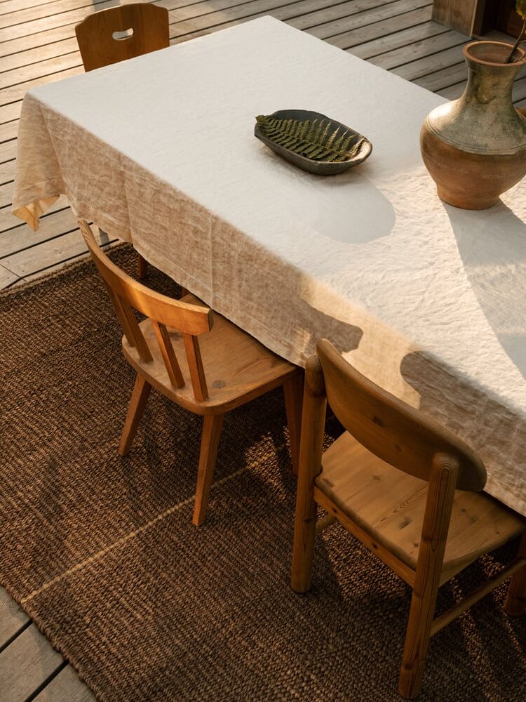 A wooden table with a white tablecloth, two wooden chairs, a ceramic vase, and a decorative dish sits on a brown woven rug on a wooden deck.