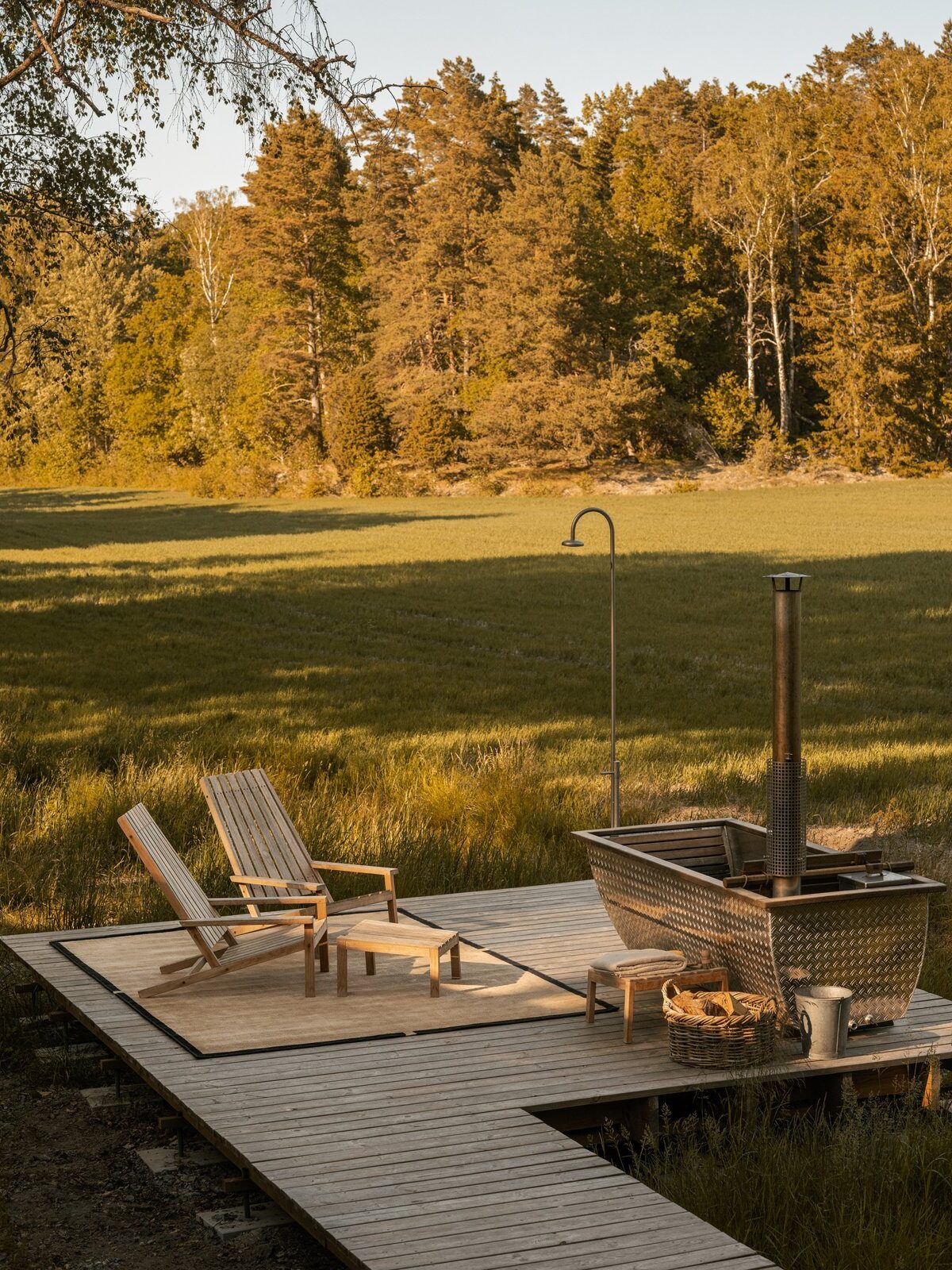 Two wooden lounge chairs and a metal outdoor hot tub are set on a wooden deck overlooking a grassy field and trees in the background.