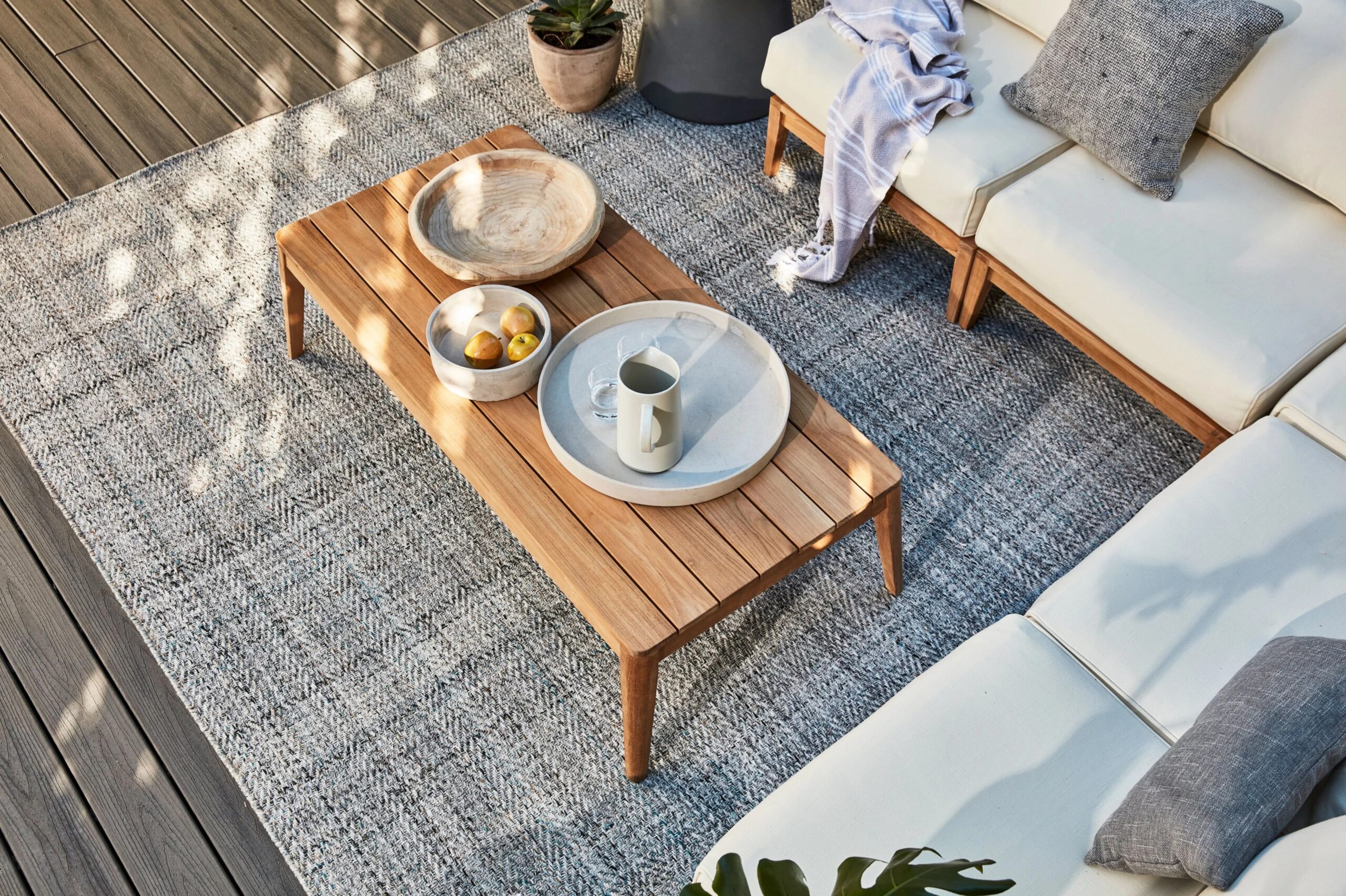 Wooden coffee table with dishes and fruit sits on a textured rug next to a white outdoor sofa with cushions and a throw blanket on a wooden deck.