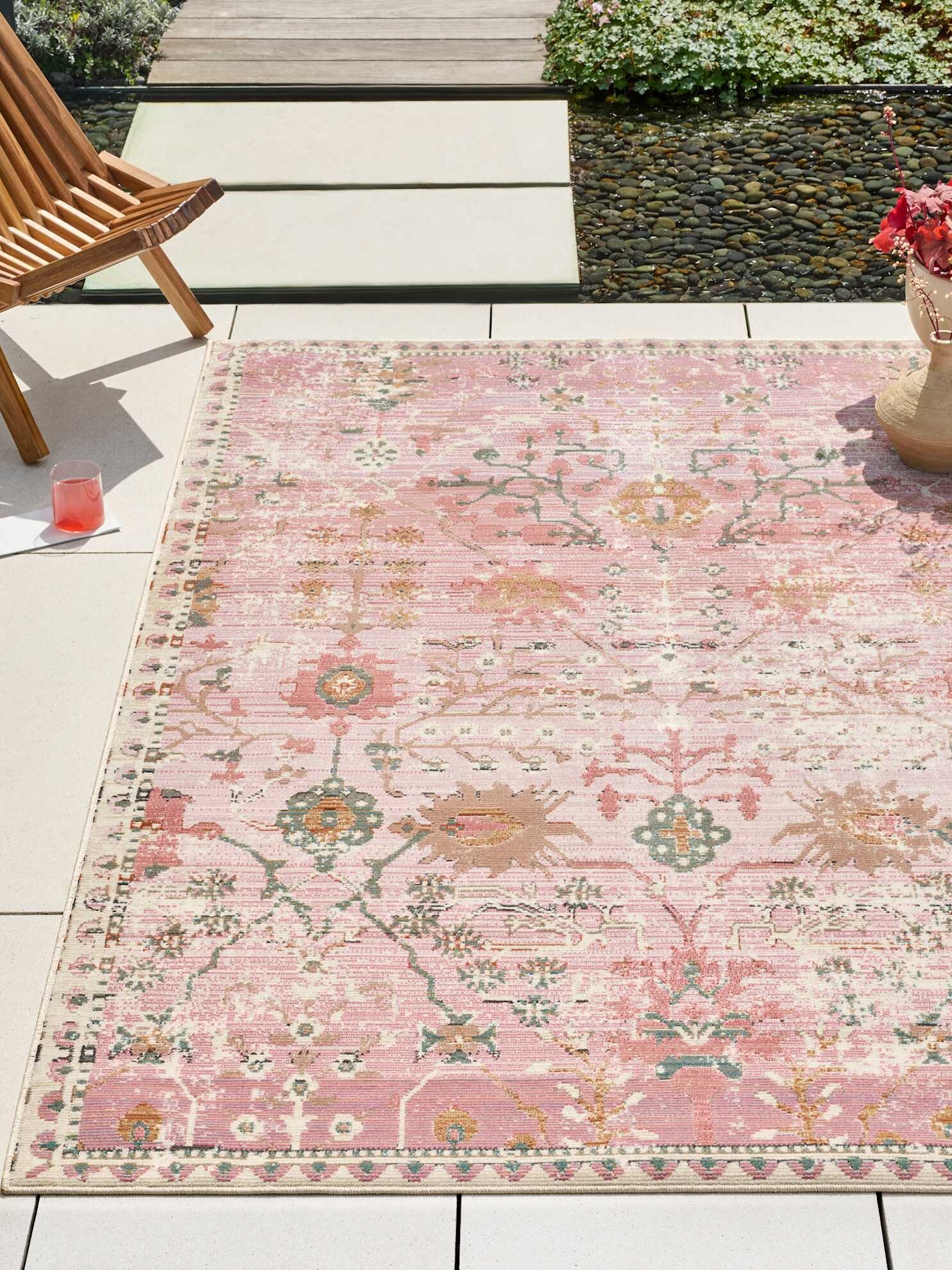 A pink patterned rug lies on a light patio near a wooden chair, a red drink, and a vase with red flowers, adjacent to a small pond and greenery.
