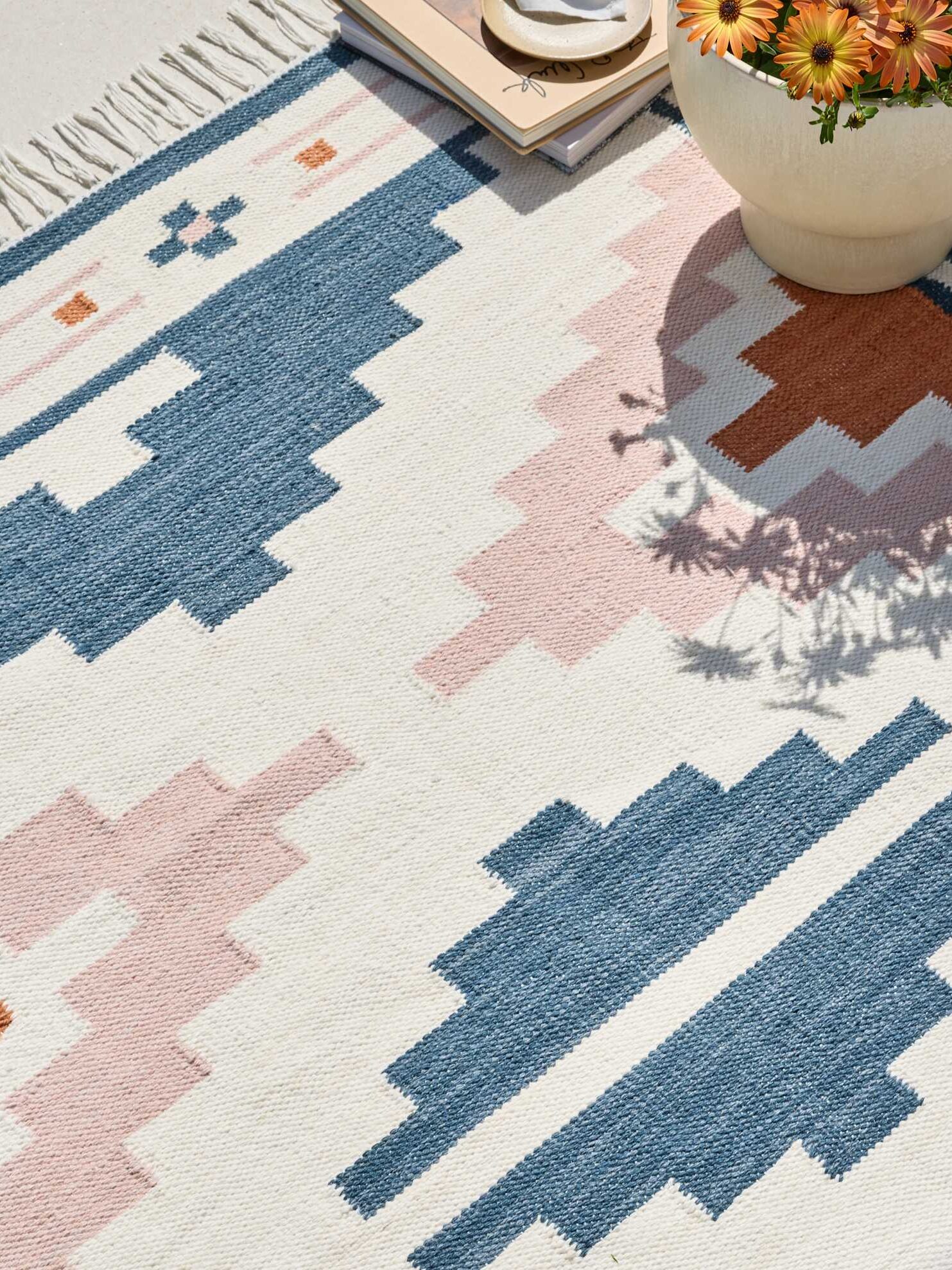 A geometric patterned rug in shades of blue, blush, and orange lies on the floor near a beige pot with flowers and a stack of books.