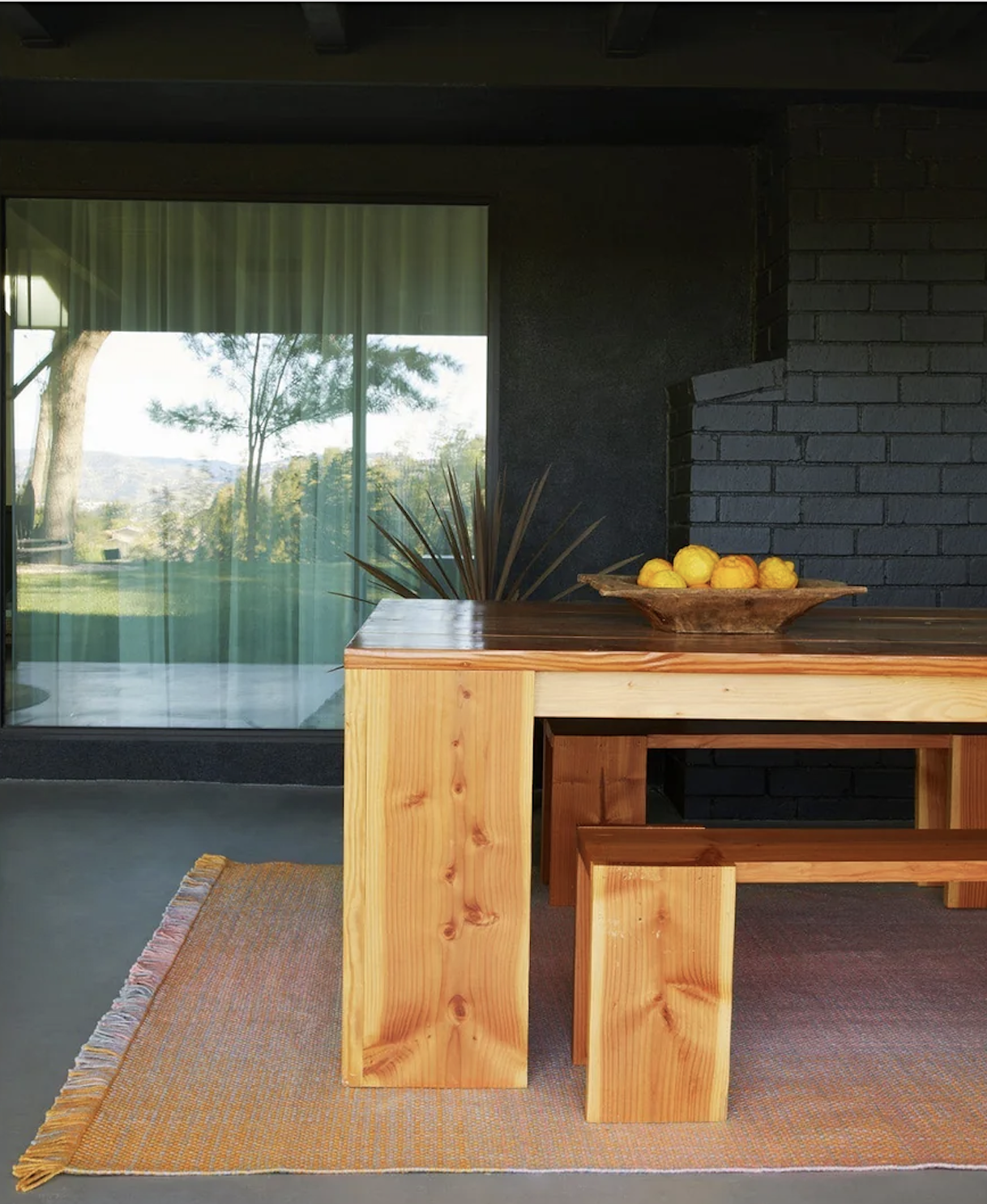 A wooden table with benches sits on a woven rug; a bowl of lemons is on the table. A large window and dark brick wall are in the background.