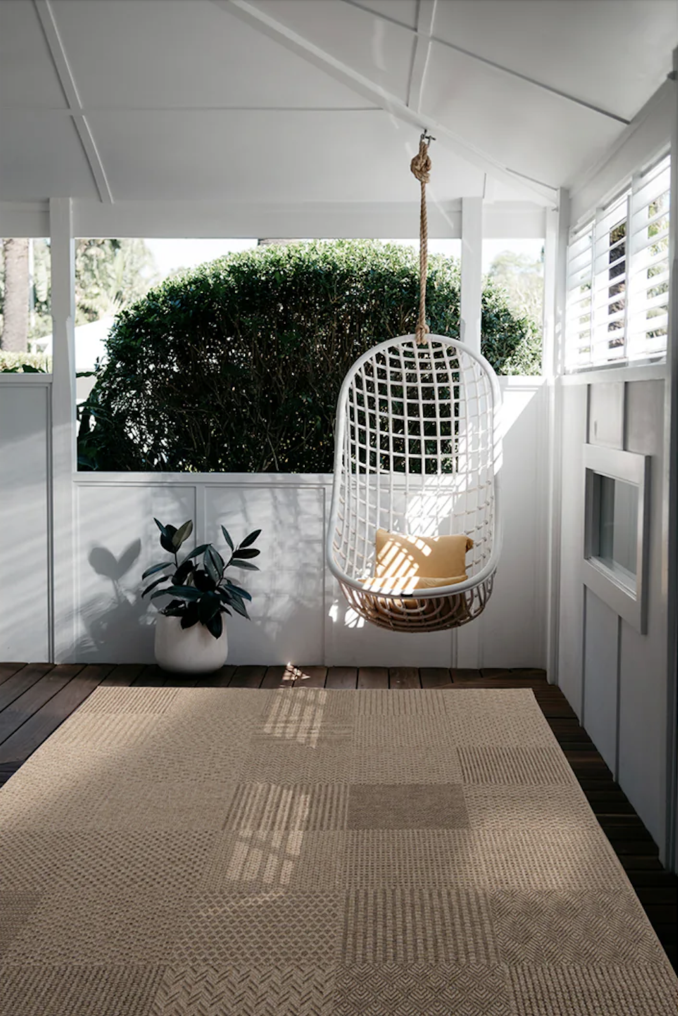 A white hanging chair with yellow cushions is suspended on a covered porch next to a potted plant and a large bush outside. Sunlight casts shadows on the floor.