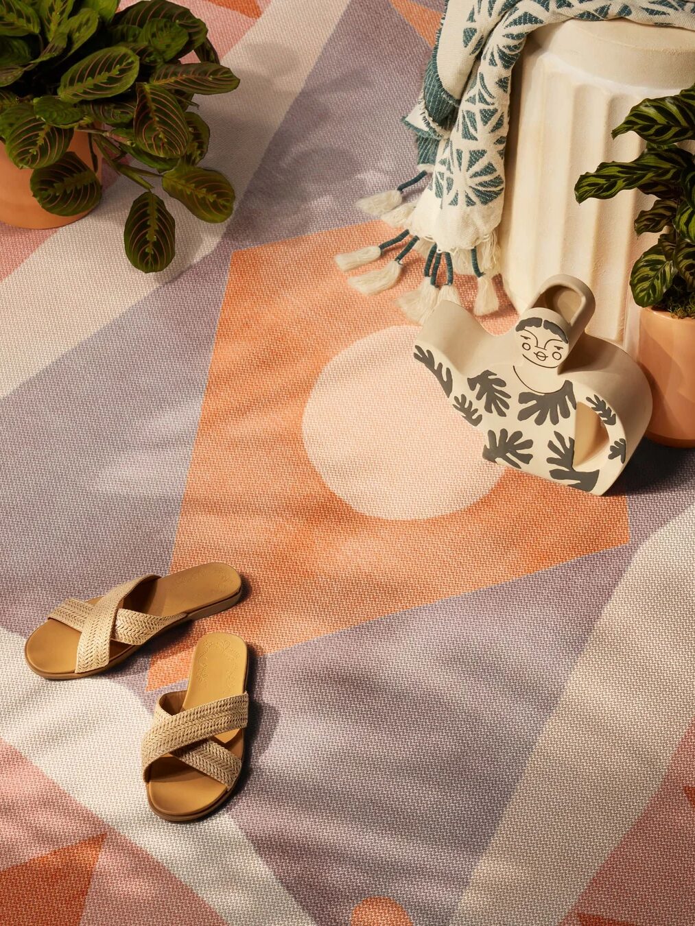 A pair of woven sandals on a geometric-patterned rug, surrounded by potted plants, a ceramic pitcher, and a draped blanket.