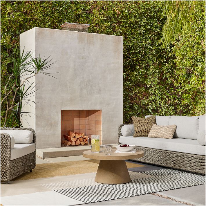 Modern outdoor patio with a concrete fireplace, beige cushioned sofas, a round coffee table with snacks, and a lush green hedge in the background.