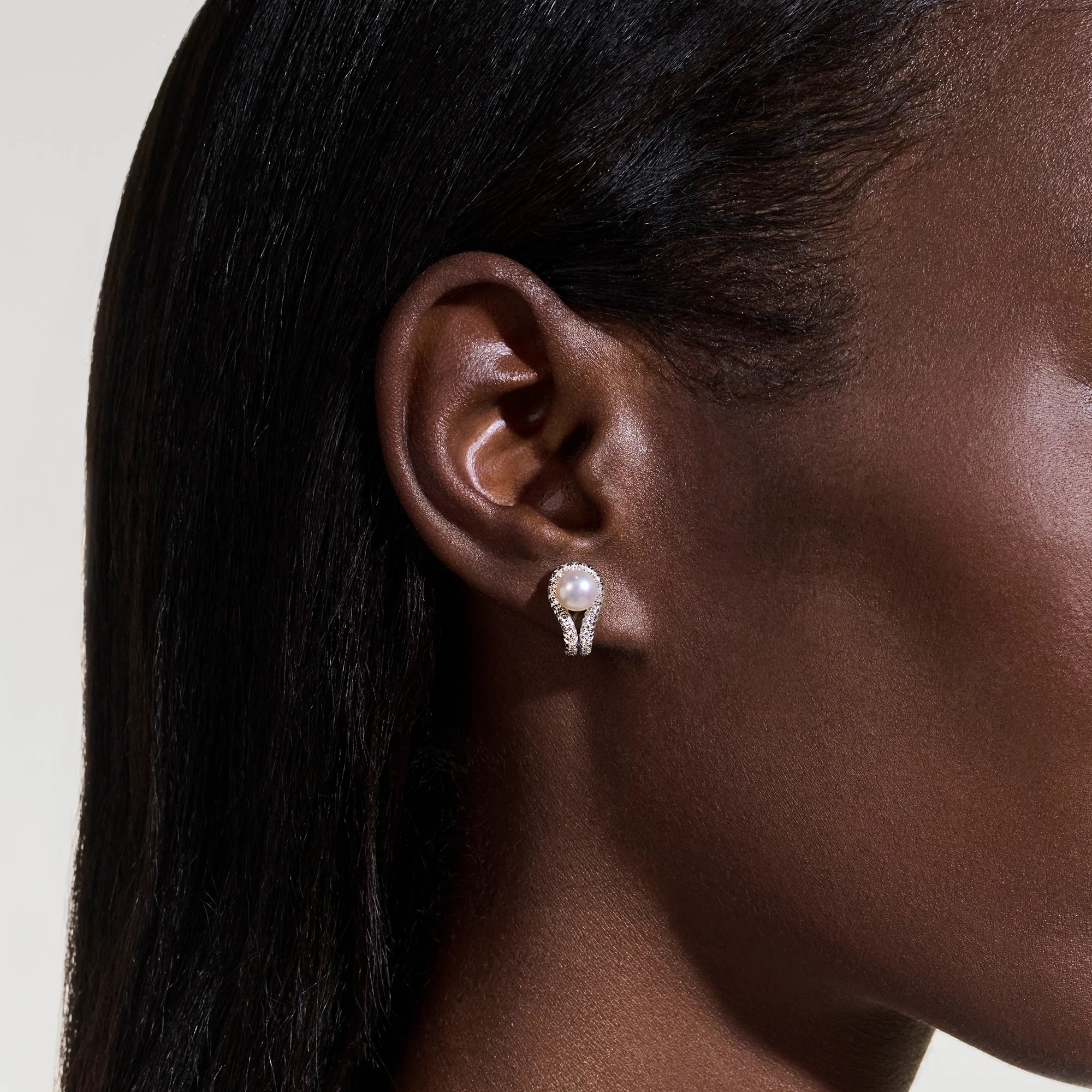 A close-up of a woman's ear wearing a pearl and diamond earring, with smooth brown skin and straight dark hair visible.