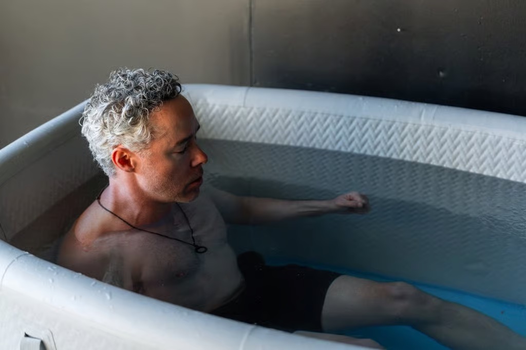 A man with curly gray hair sits waist-deep in an inflatable tub of water, eyes closed, and wearing black shorts and a necklace.
