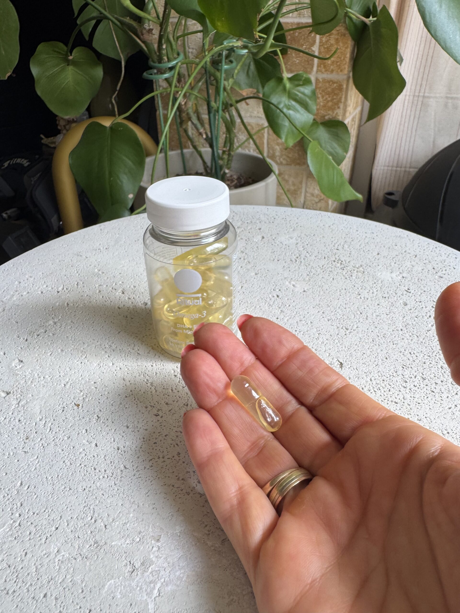 A hand holding two clear capsules in front of a bottle of similar capsules on a white table, with green plants in the background.