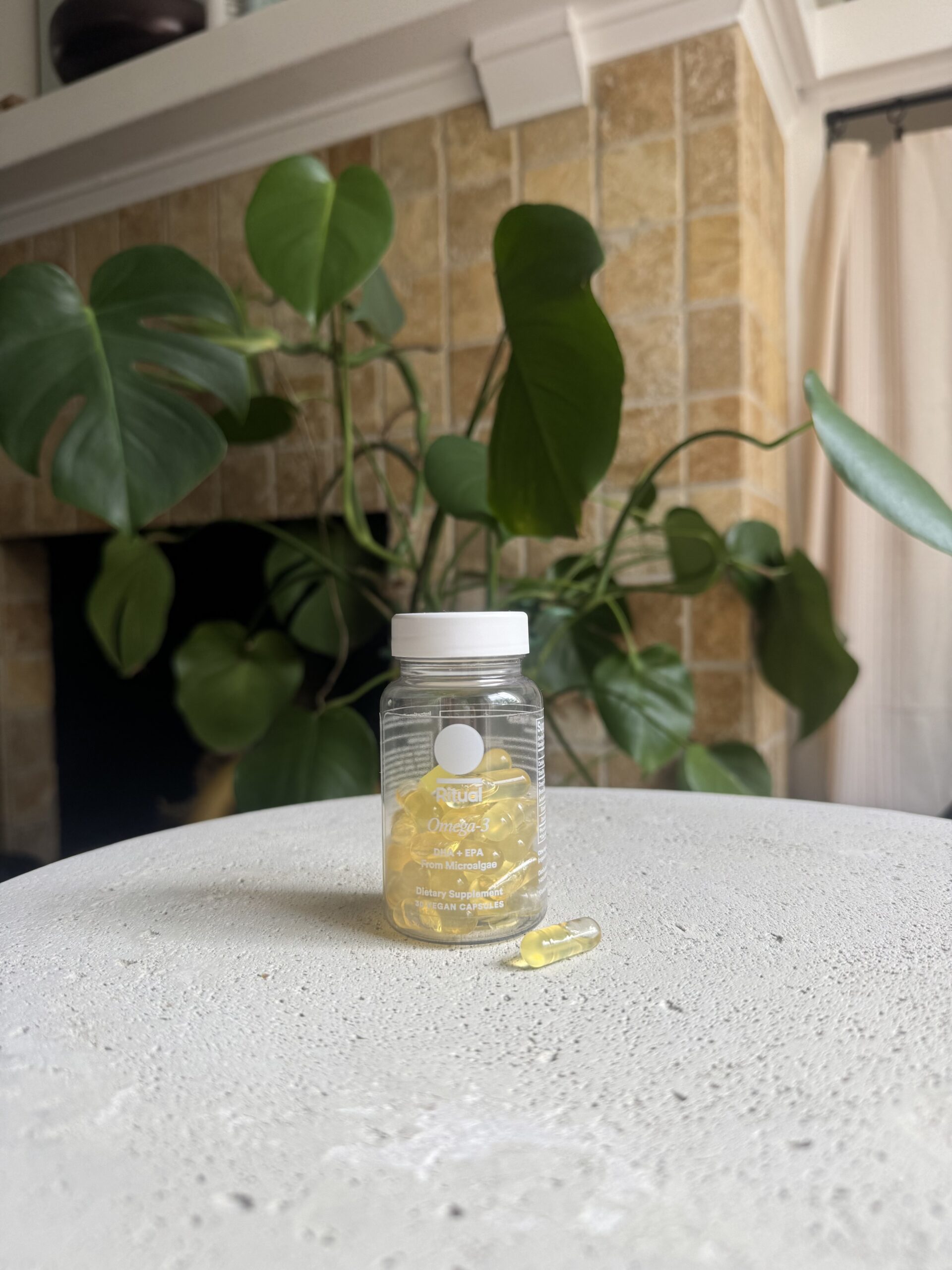 A clear bottle of Omega-3 fish oil capsules sits on a light-colored surface, with one capsule lying next to it; green plants and a tiled wall are in the background.