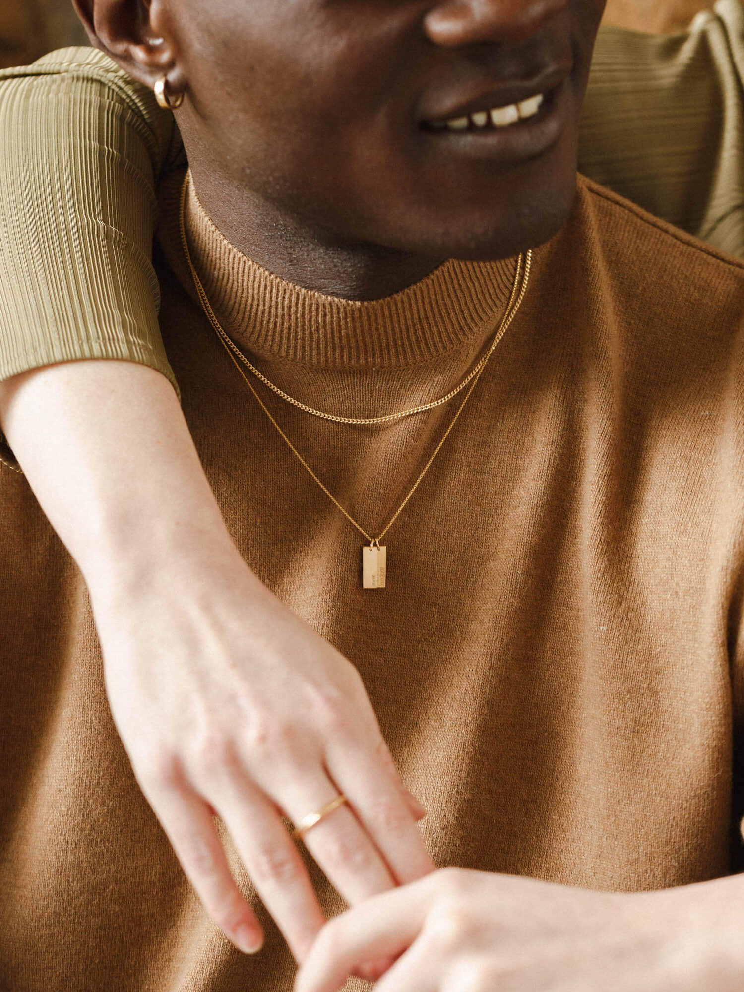 A person in a brown sweater with gold necklaces is embraced by another person with a gold ring, both partially visible.