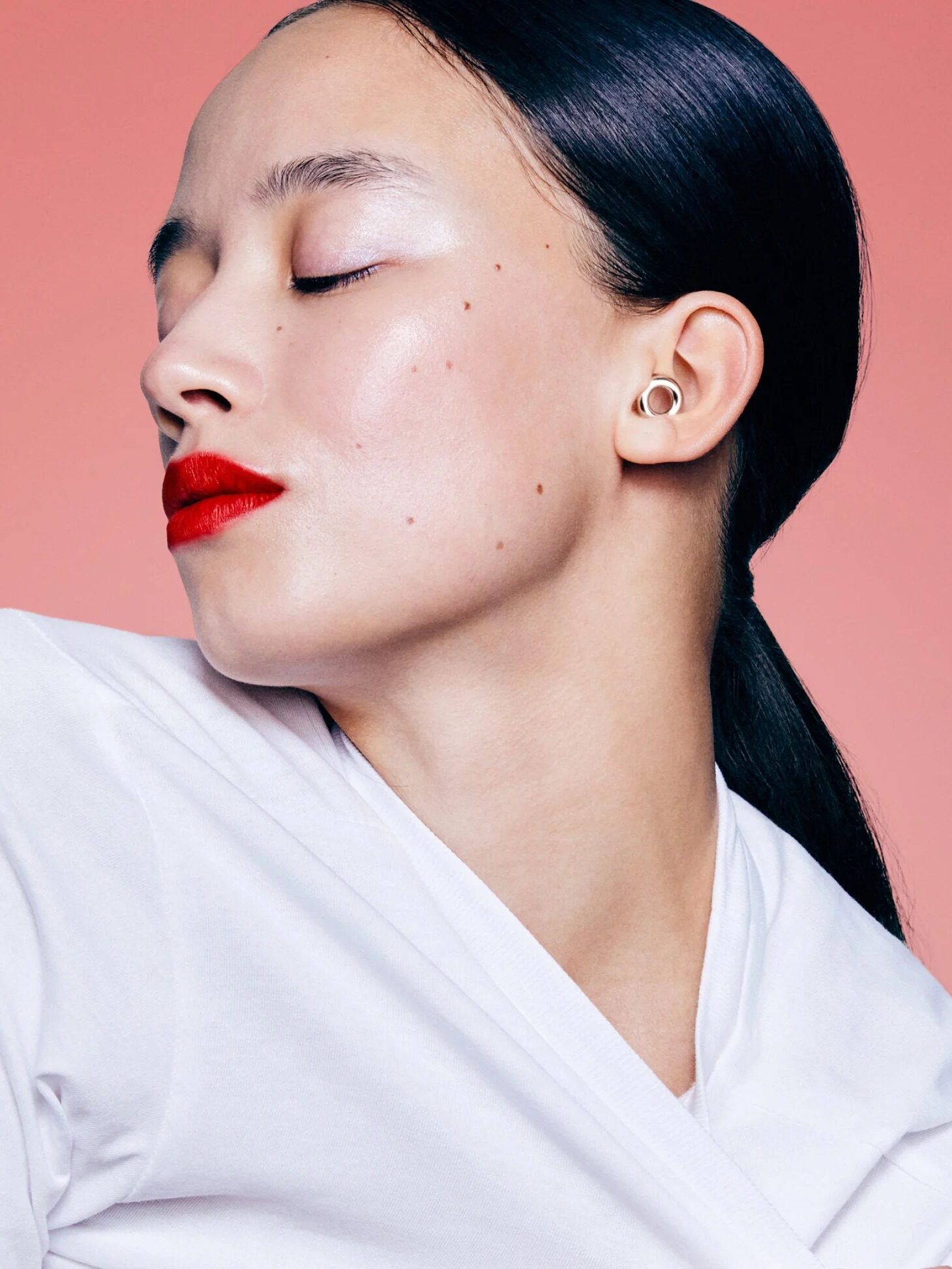 Woman with dark hair in a low ponytail poses against a pink background, wearing a white top, red lipstick, and small stud earrings, with her eyes closed.