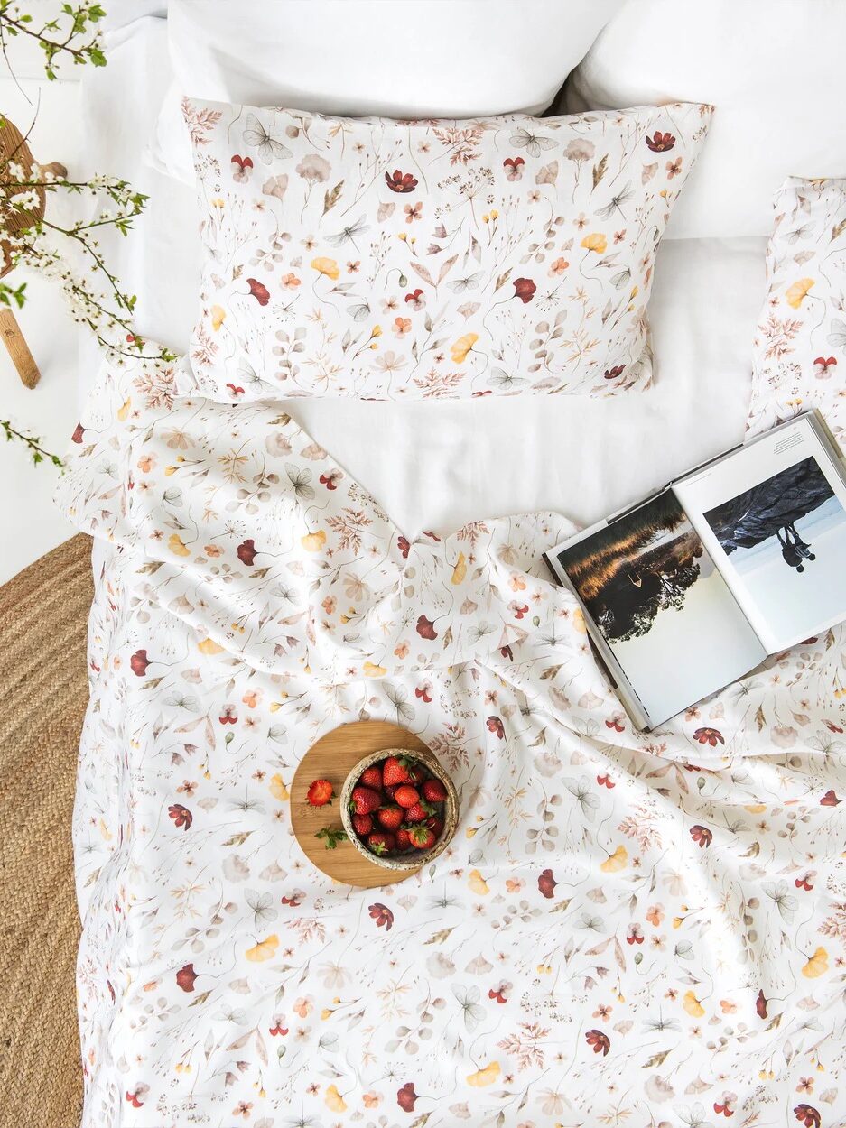 A bed with floral-patterned sheets, a pillow, an open magazine, and a wooden bowl of strawberries on top, next to a round woven rug and a vase with branches.