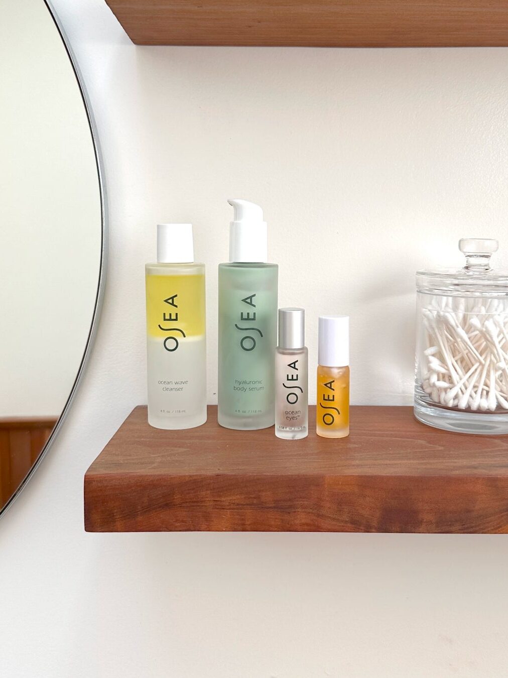 A wooden shelf holds four OSEA skincare products and a glass jar filled with cotton swabs, next to a round mirror on a white bathroom wall.