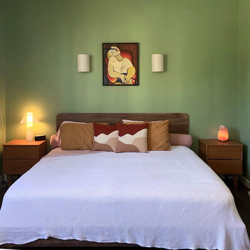 A neatly made bed with three pillows and a white cover, flanked by two nightstands with lamps, against a green wall with a framed abstract painting above the headboard.