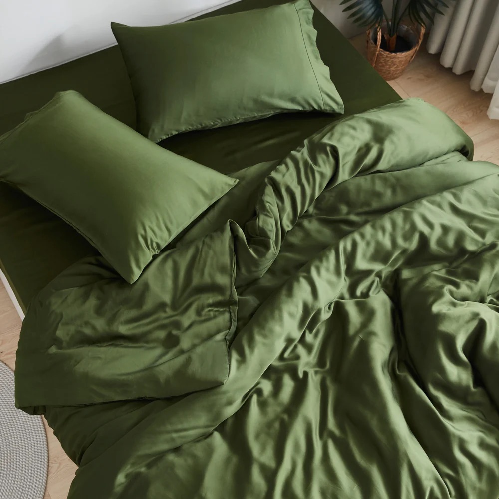 Green bedding set with two pillows and a comforter on a bed, slightly rumpled, in a minimally decorated room with a plant visible in the corner.