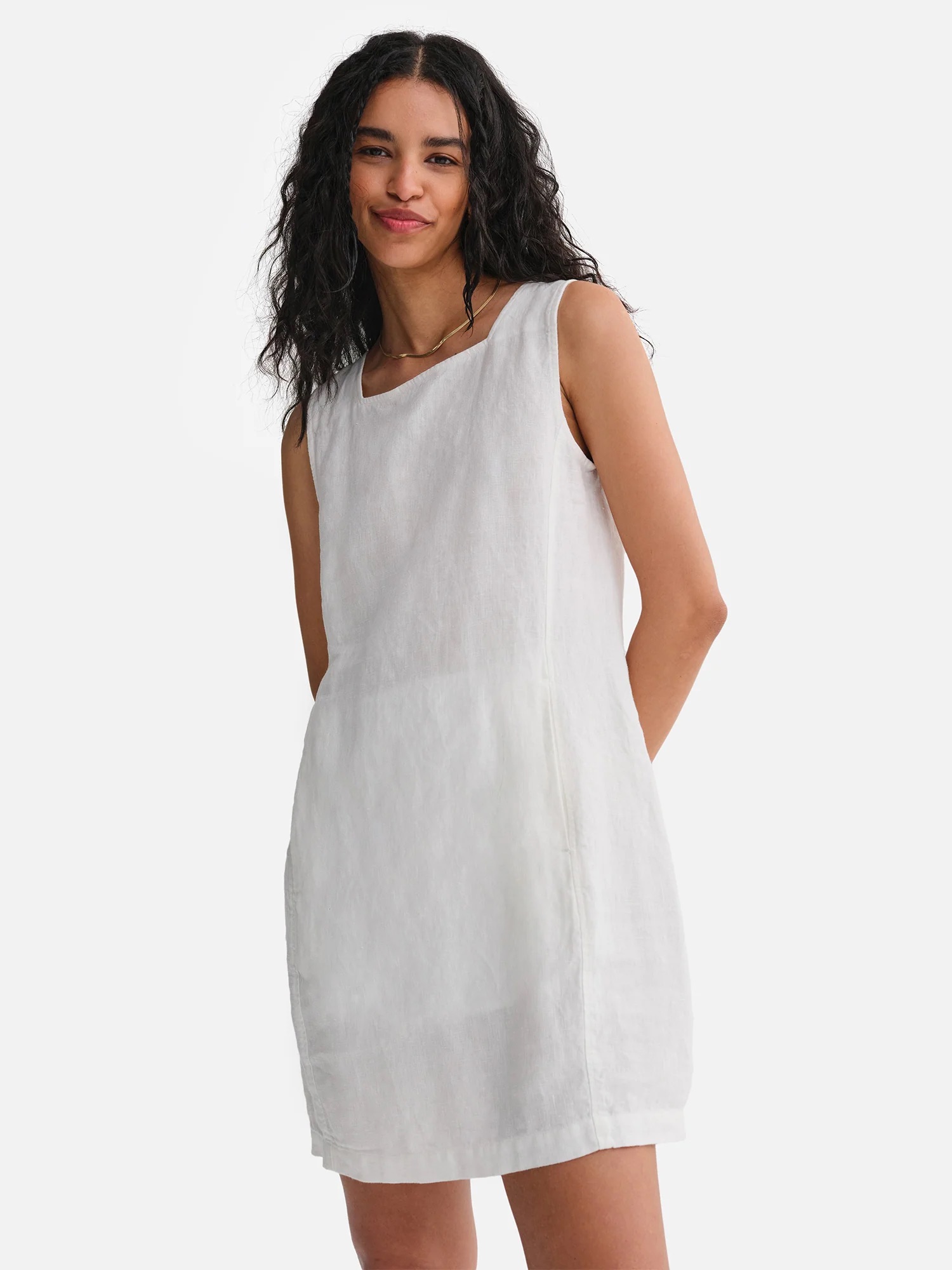A woman with long curly hair wears a sleeveless, knee-length white linen dress and stands against a plain white background.