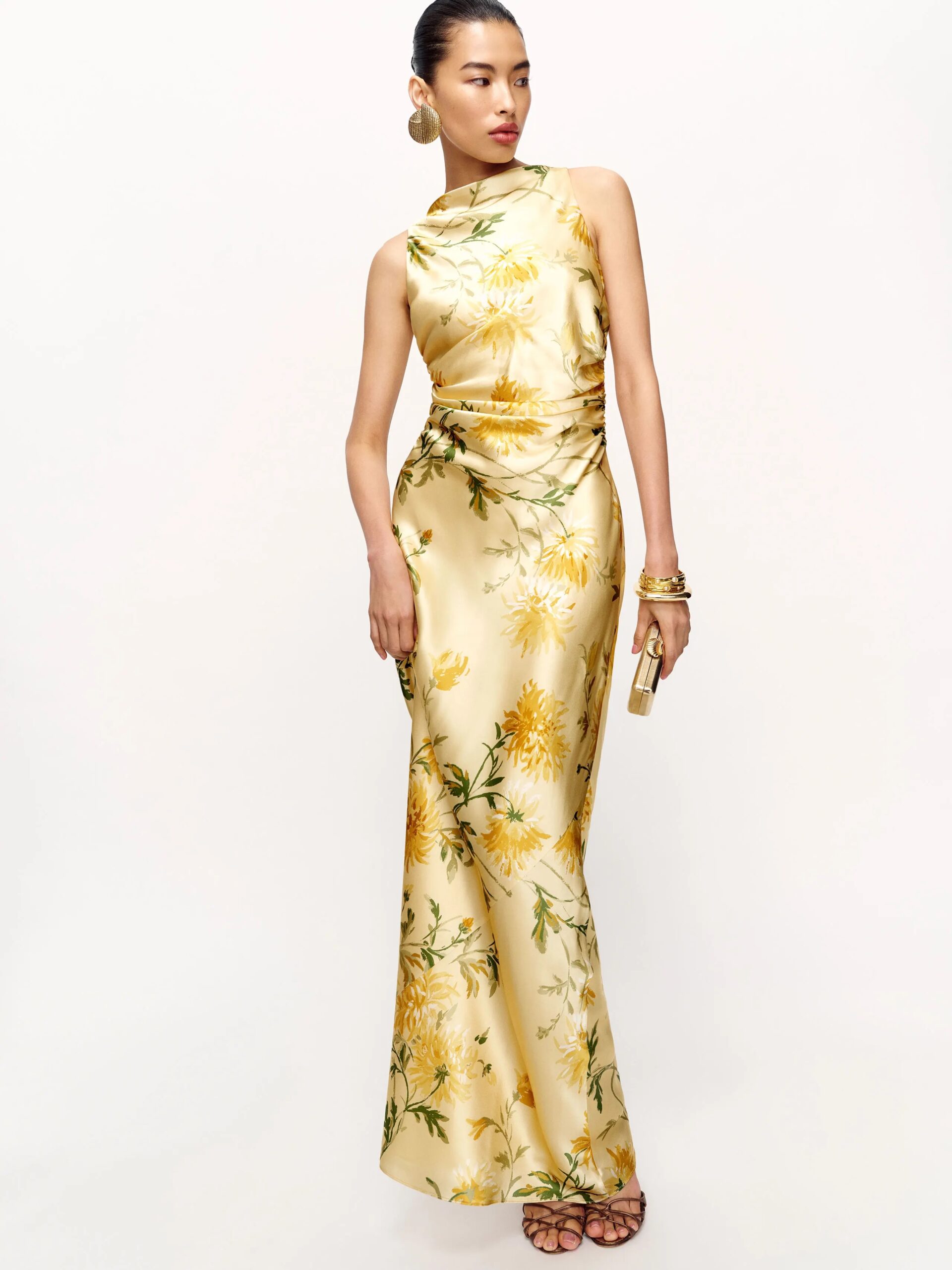 A woman stands wearing a sleeveless, floor-length, yellow floral silk dress with gold accessories and high-heeled sandals against a plain white background.