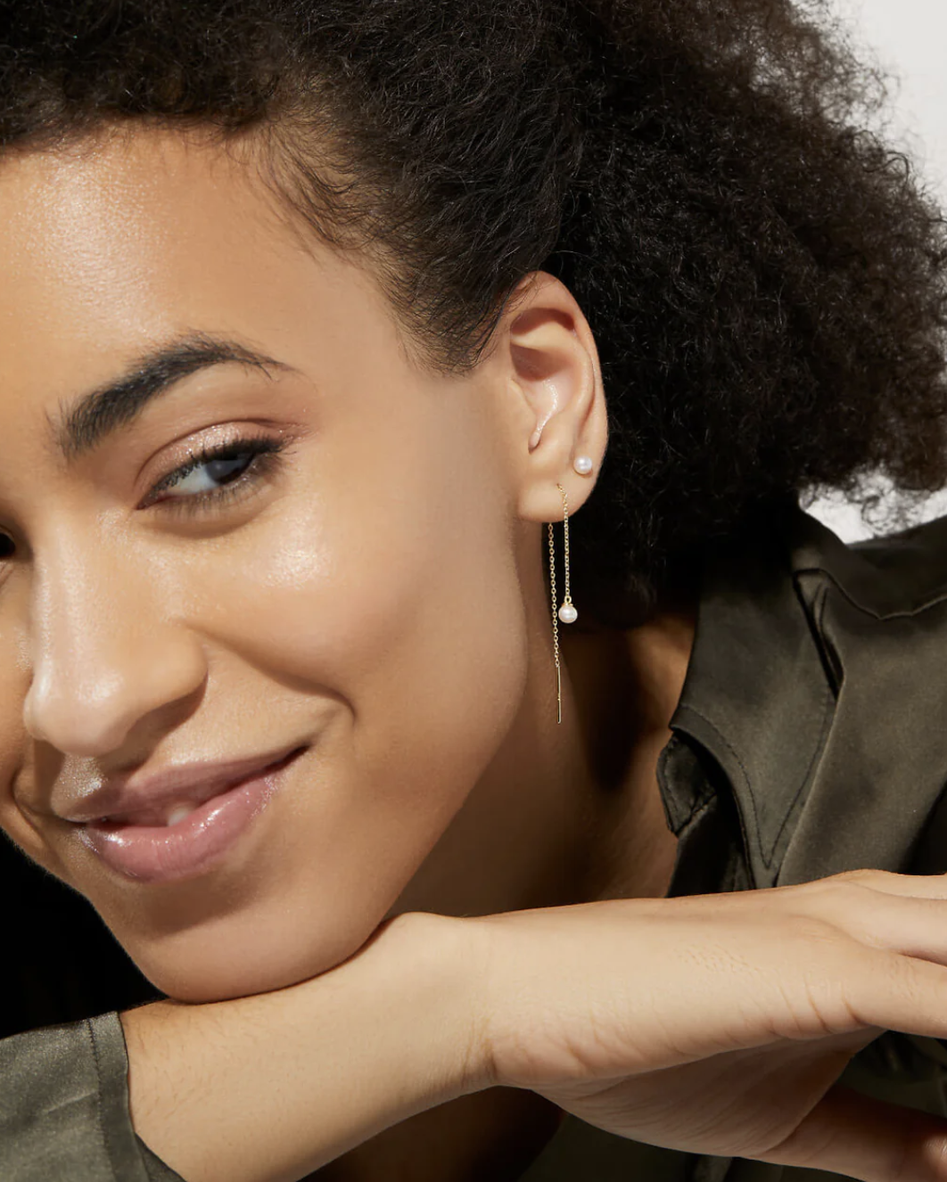 A woman with curly hair smiles, resting her chin on her hands, and wears a gold dangle earring with a small pearl.
