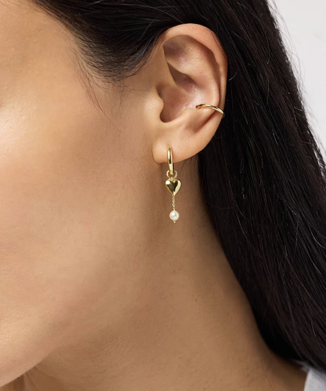 A close-up of a woman's ear wearing a gold hoop earring with a heart-shaped charm and a dangling pearl, and a simple gold ear cuff on the upper ear.