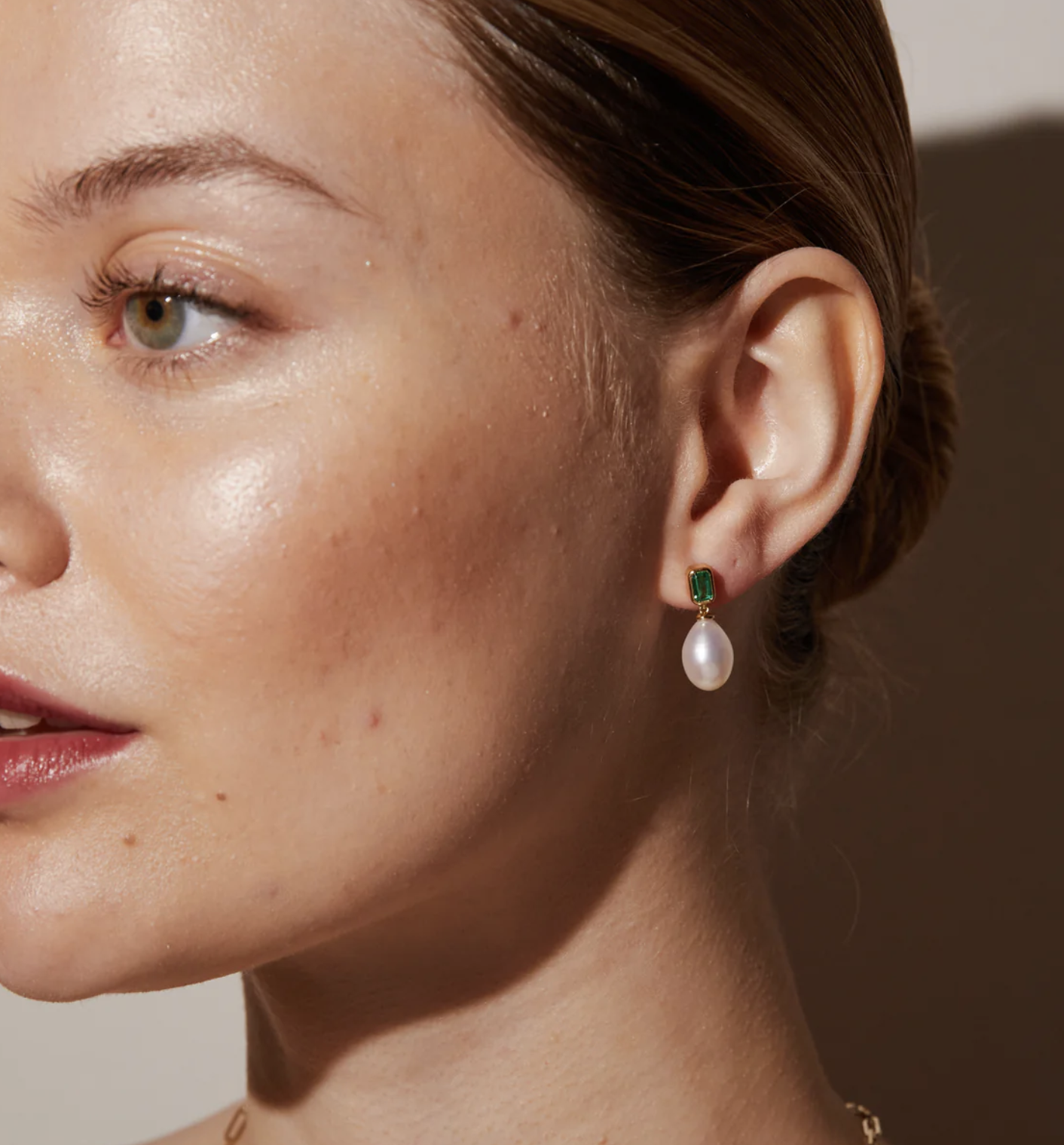 Close-up of a woman wearing an earring with a green gemstone stud and a dangling white pearl, against a neutral background.