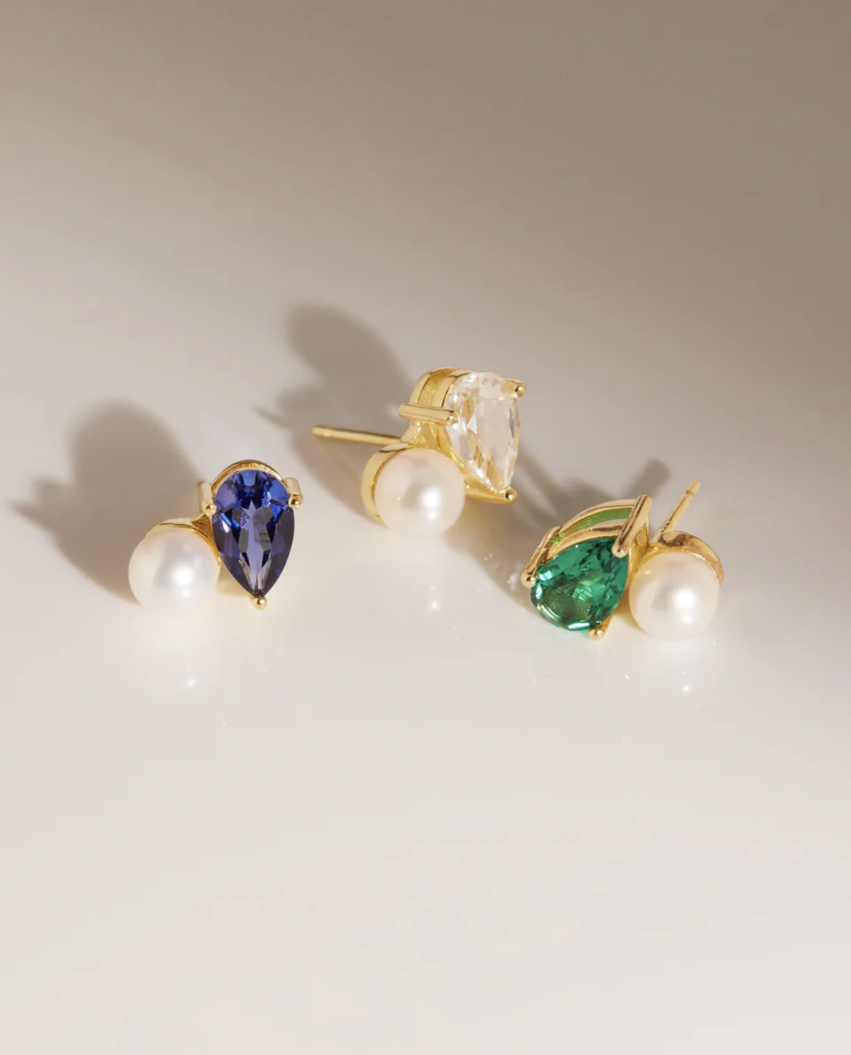 Three gold stud earrings are displayed, each featuring a pearl and a teardrop-shaped gemstone—one blue, one clear, and one green—on a neutral surface.