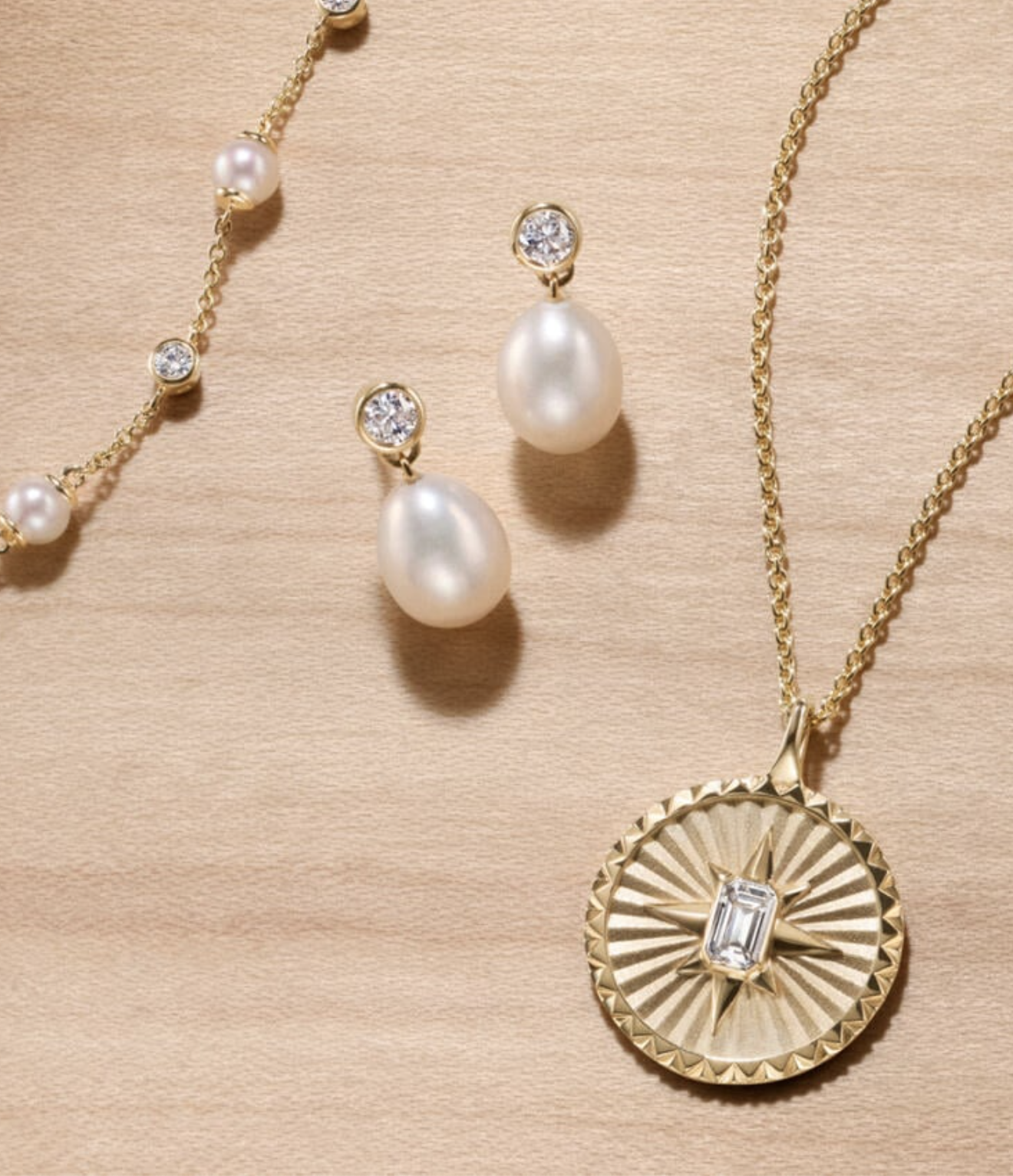Gold necklace with a round, textured pendant, matching chain, a pair of pearl and diamond drop earrings, and a gold bracelet with pearls, all displayed on a wooden surface.