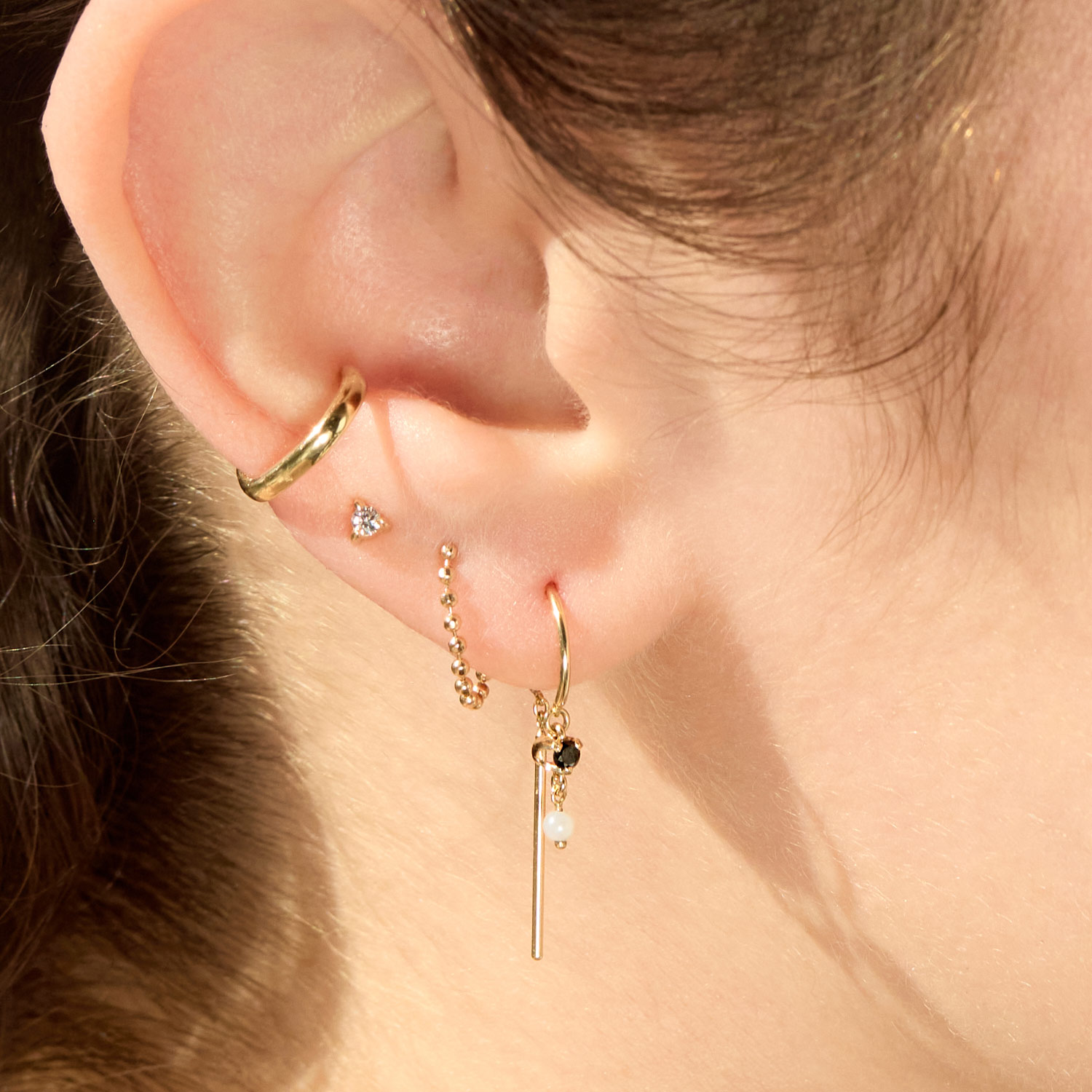 Close-up of an ear with multiple gold earrings, including a cuff, a heart stud, a chain stud, and a dangling bar earring with black and white beads.