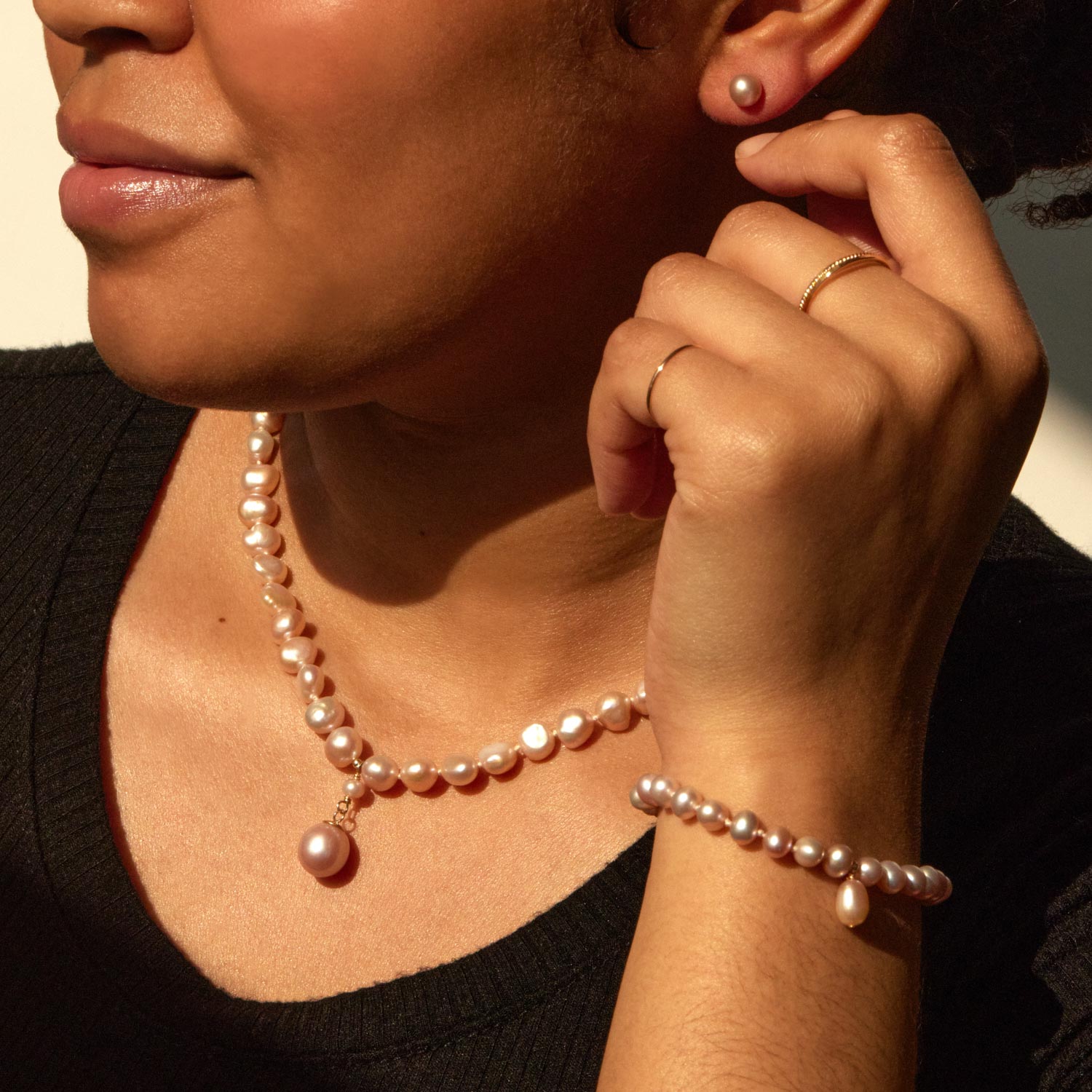 A woman wearing a pearl necklace with a pendant, a matching pearl bracelet, a pearl stud earring, and two thin gold rings on her fingers.