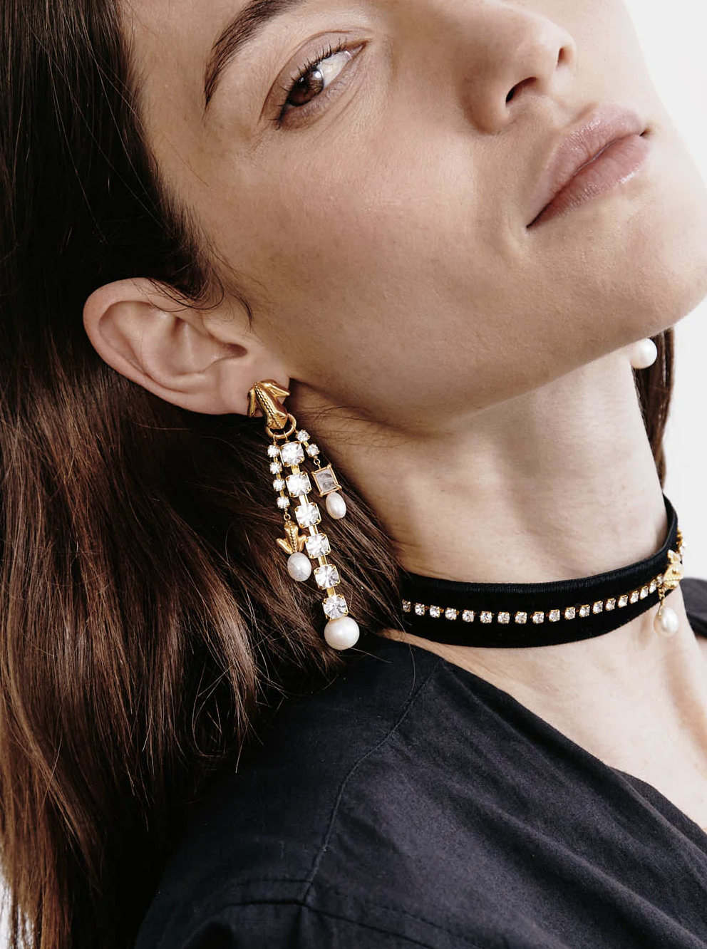 A woman wearing a gold and pearl chandelier earring and a black choker necklace with pearl accents, shown from the shoulders up.