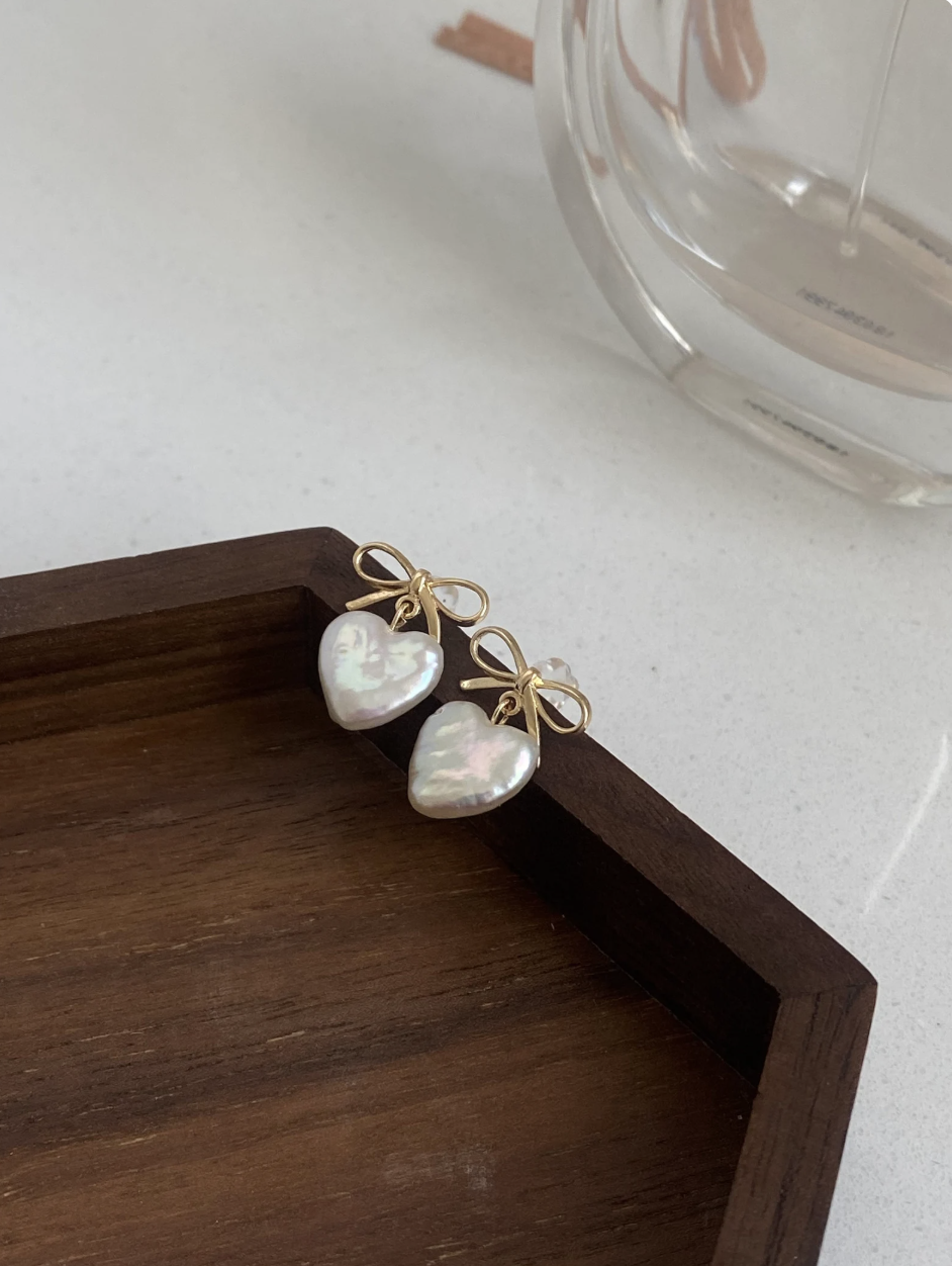 A pair of gold earrings with heart-shaped mother-of-pearl pendants and small bow accents, resting on a wooden tray.
