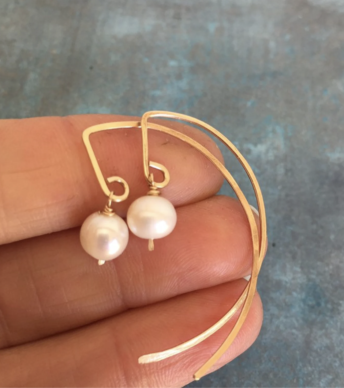 A hand holding a pair of gold wire earrings with white pearl drops against a blue textured background.
