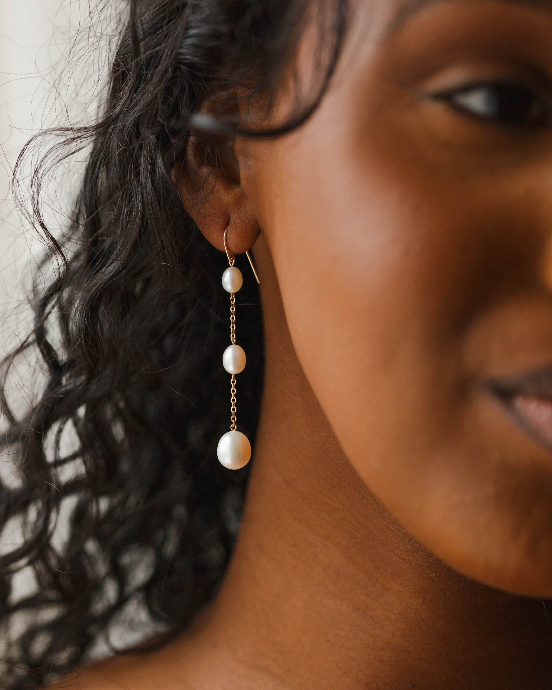 Close-up of a person wearing a gold chain earring with three white pearls of varying sizes, against a blurred background.