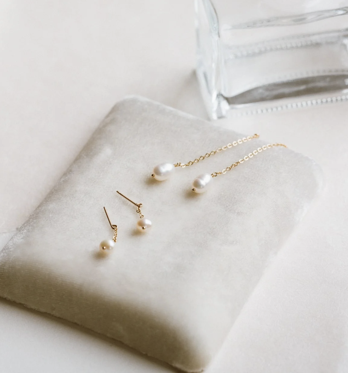 A pair of gold pearl drop earrings and a matching gold pearl necklace are displayed on a white velvet cushion next to a clear glass object.