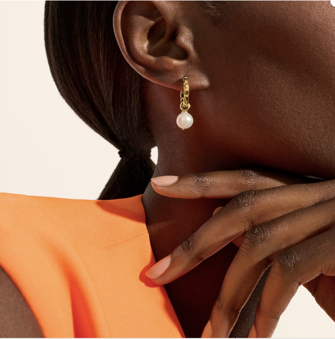 A person wearing a gold hoop earring with a pearl drop, dressed in an orange top, rests their hand on their chin.