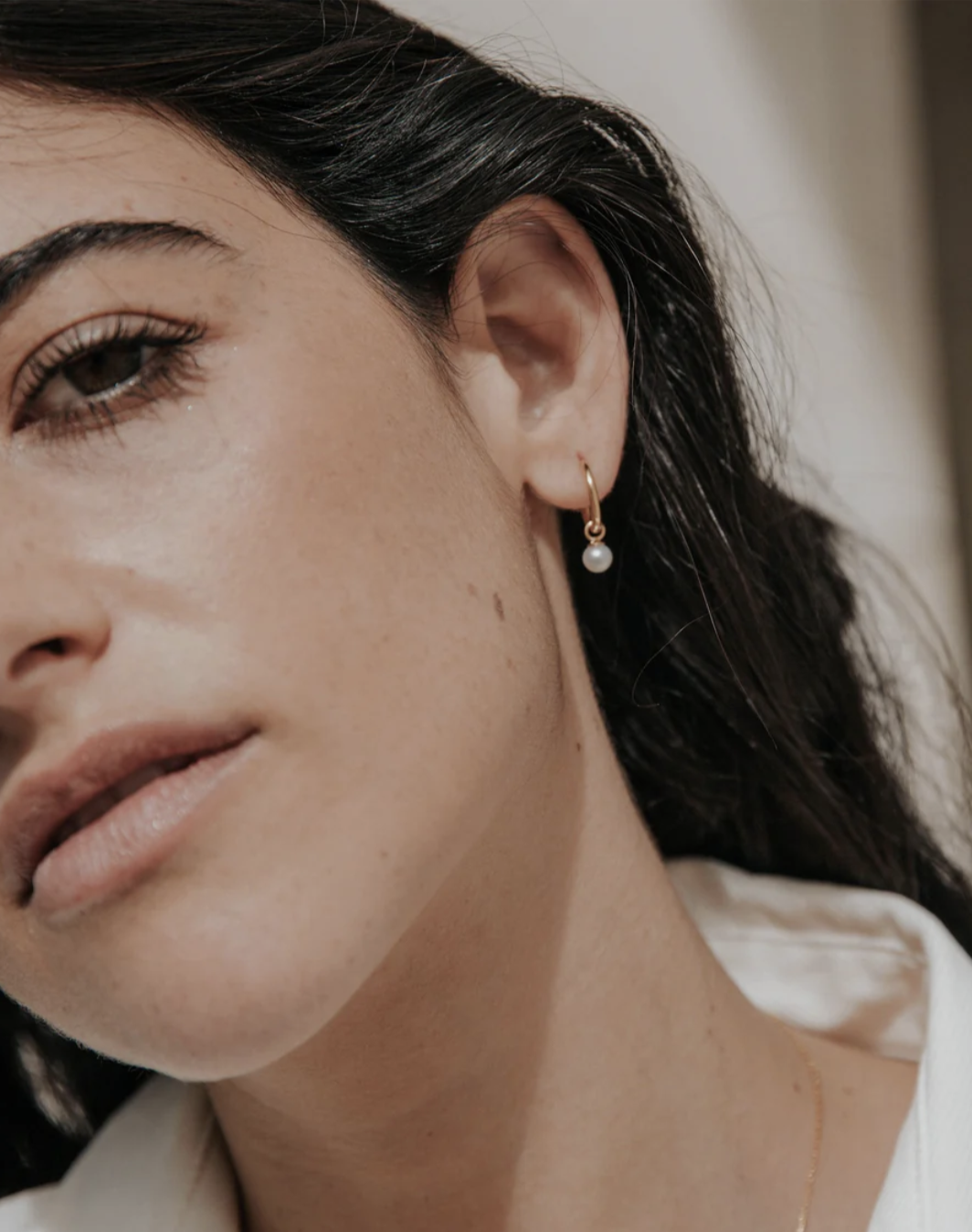 Close-up of a woman's face, showing a gold hoop earring with a small pearl. She has dark hair, natural makeup, and is wearing a white collared shirt.