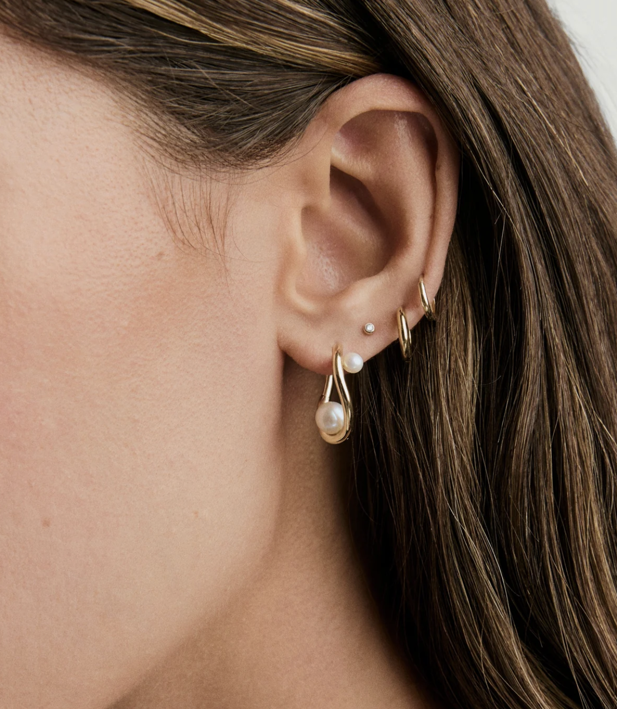 Close-up of a woman's ear with five piercings, showcasing gold hoop earrings, a pearl drop earring, and a small stud earring, with brown hair partially visible.